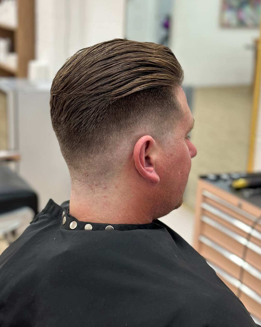 Side view of a man with a fresh, slicked-back hairstyle in a barbershop, wearing a black cape with silver buttons.
