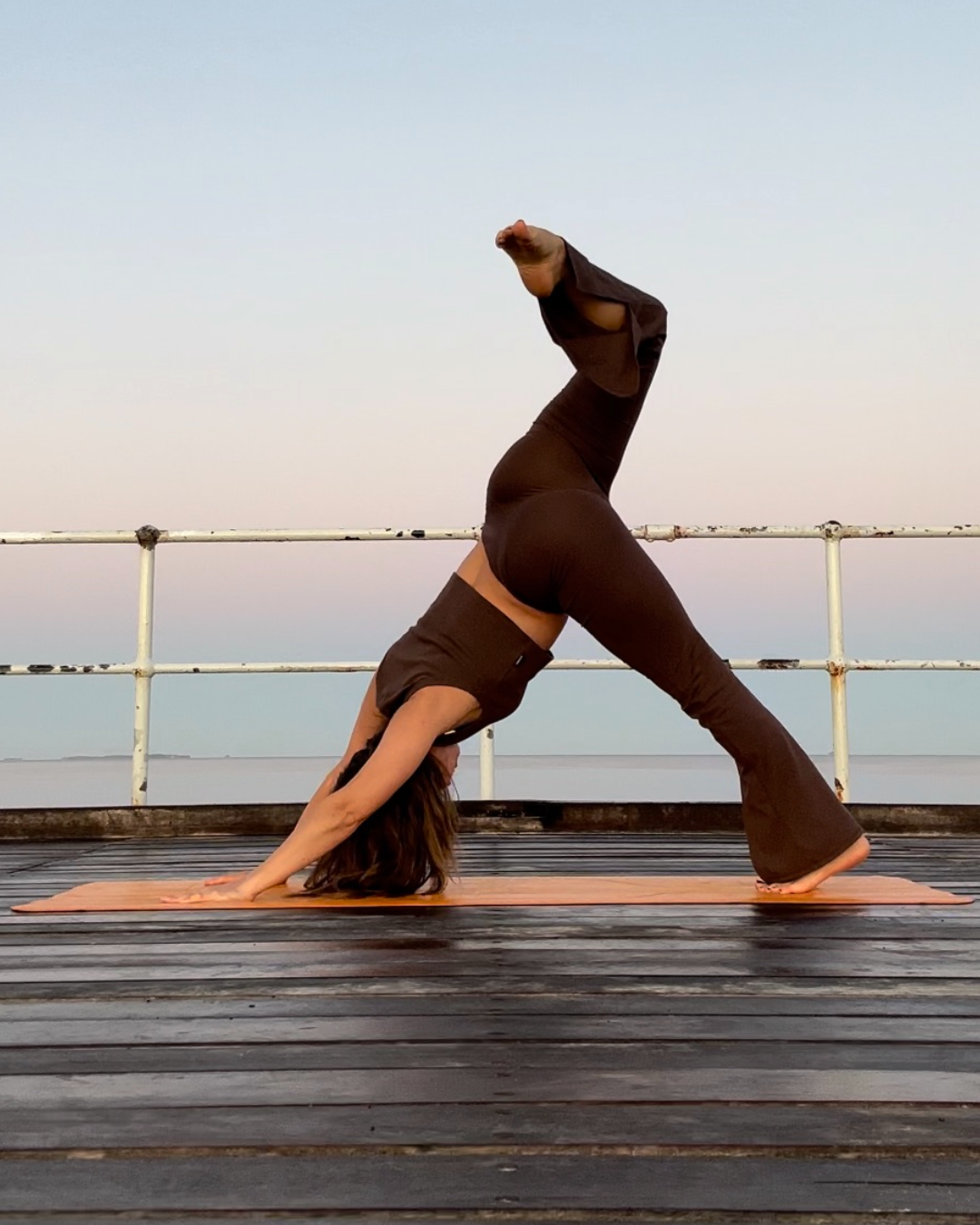 A woman practicing yoga outdoors on a wooden deck, performing a downward-facing dog pose with one leg raised, overlooking a body of water during sunset.