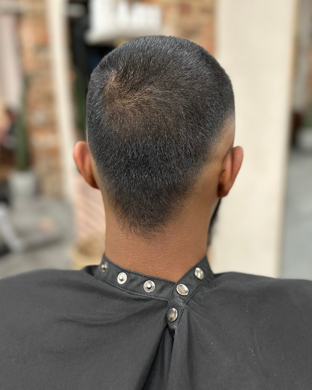 Back view of a man's head with a fresh, close-cropped hairstyle in a barbershop, wearing a black smocks with silver buttons.