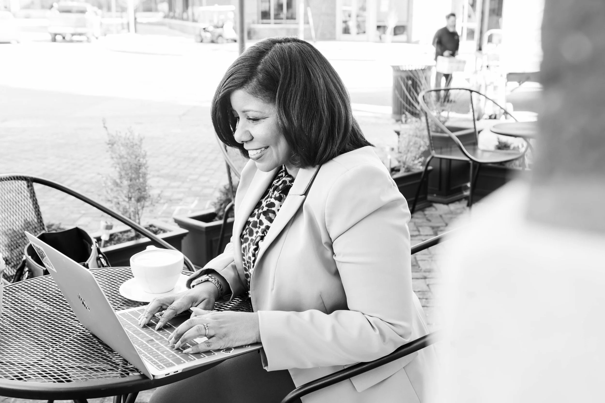 A woman with medium-length dark hair working on a laptop at an outdoor café, smiling and wearing a light-colored blazer and a leopard print scarf, with a coffee cup on the table.