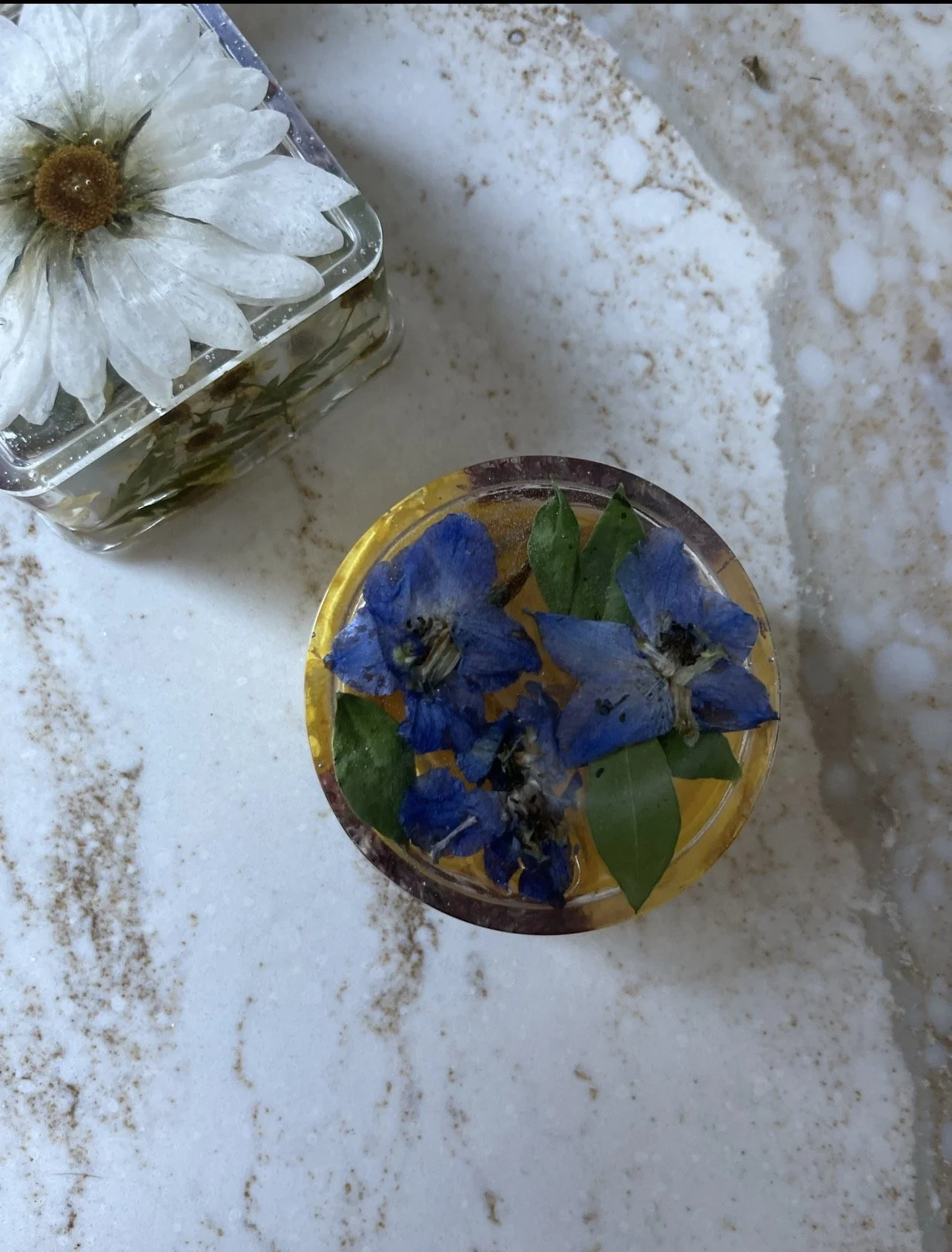 A white daisy in a clear glass vase floating in water, and a round glass container with blue flowers and green leaves inside, placed on a white and beige marble surface.