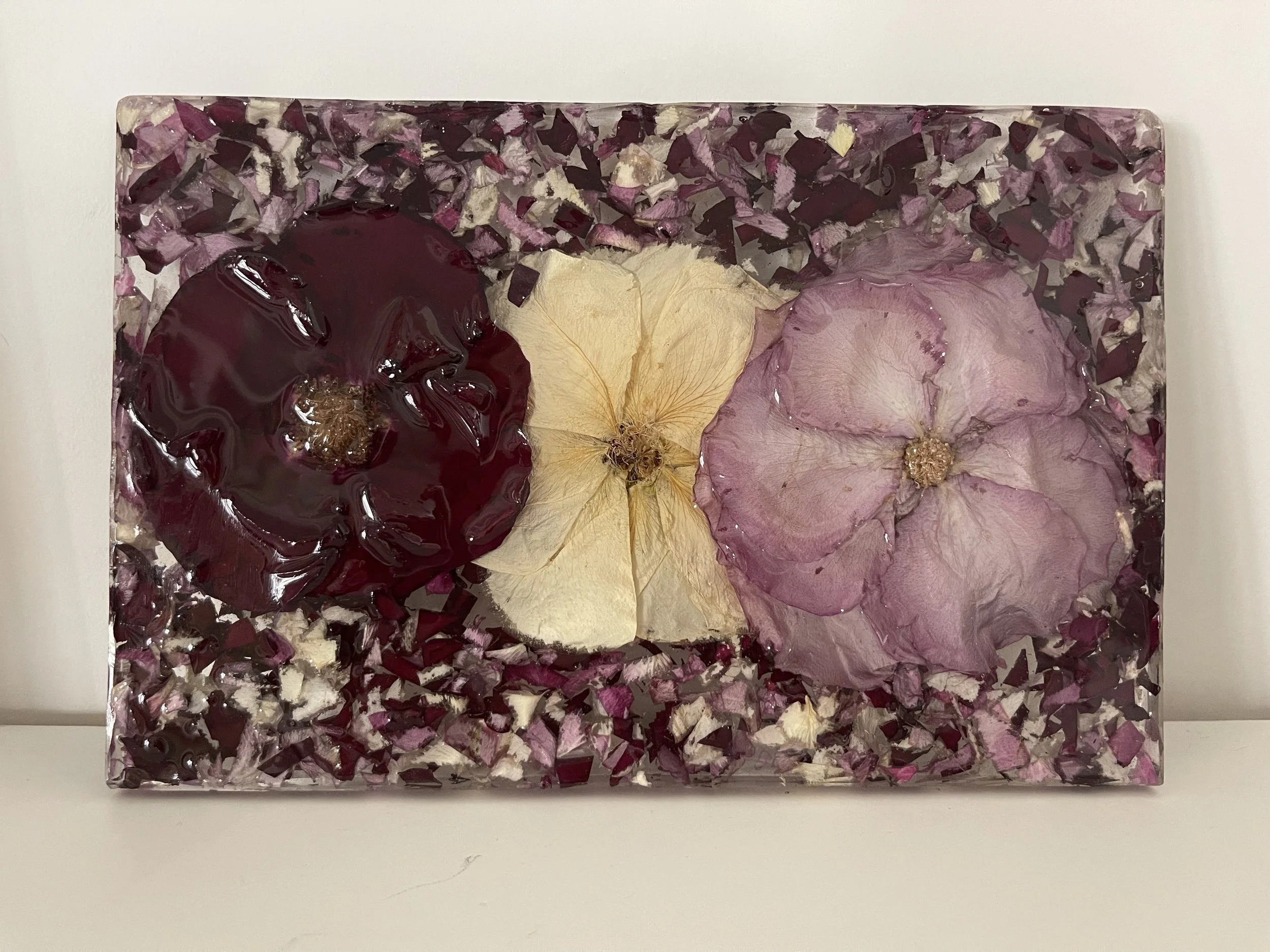 A rectangular glass art piece embedded with dried purple, beige, and pink flowers and petals, resting on a white surface against a plain white background.