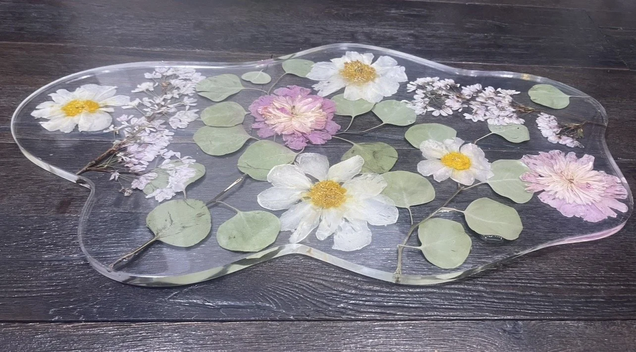 Clear decorative tray with pressed flowers and leaves on a dark wooden surface.