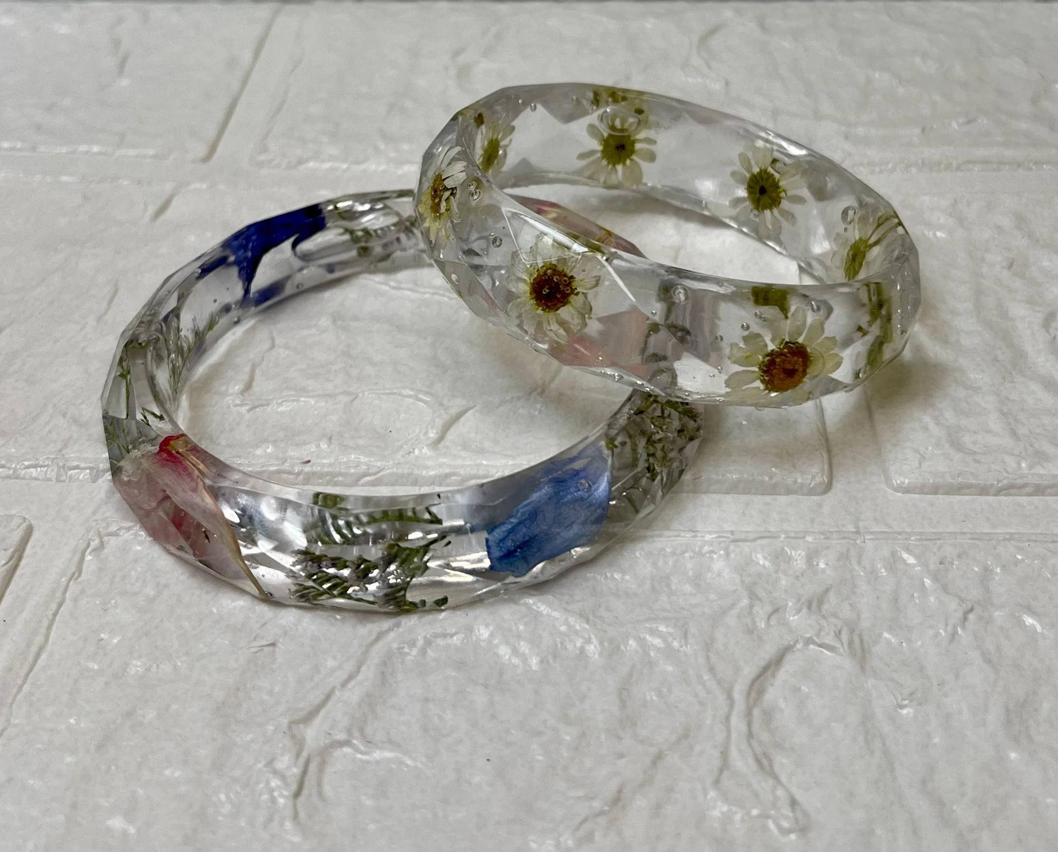 Two clear resin bracelets with embedded small dried flowers and leaves resting on a white textured surface.