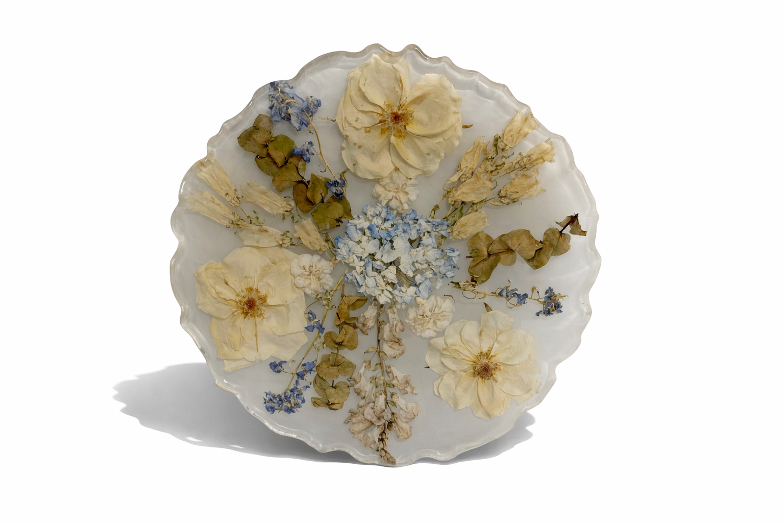 A decorative glass plate with dried flowers including cream-colored, blue, and green leaves, arranged in a pattern, casting a shadow on a white background.