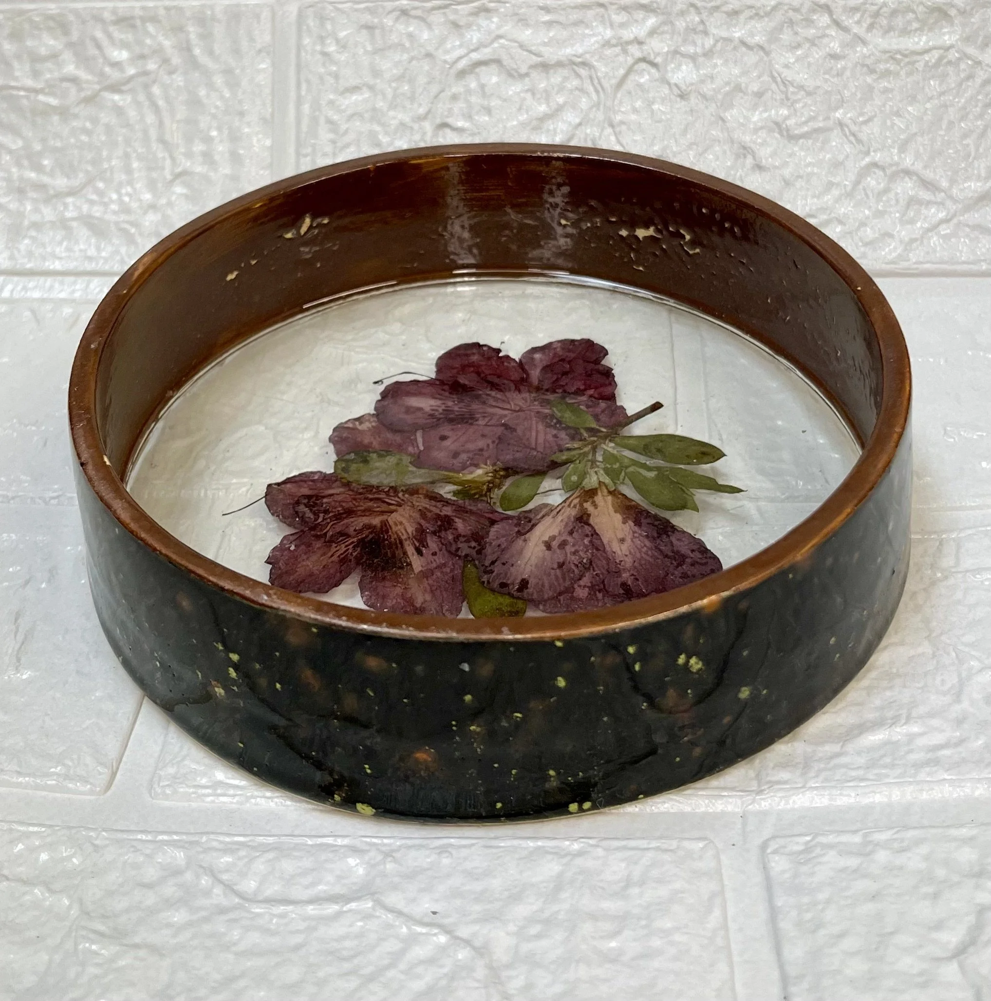 A round, dark-colored bowl with a brown rim, containing water and floating autumn leaves of various colors, including red and green, on a white tiled surface.
