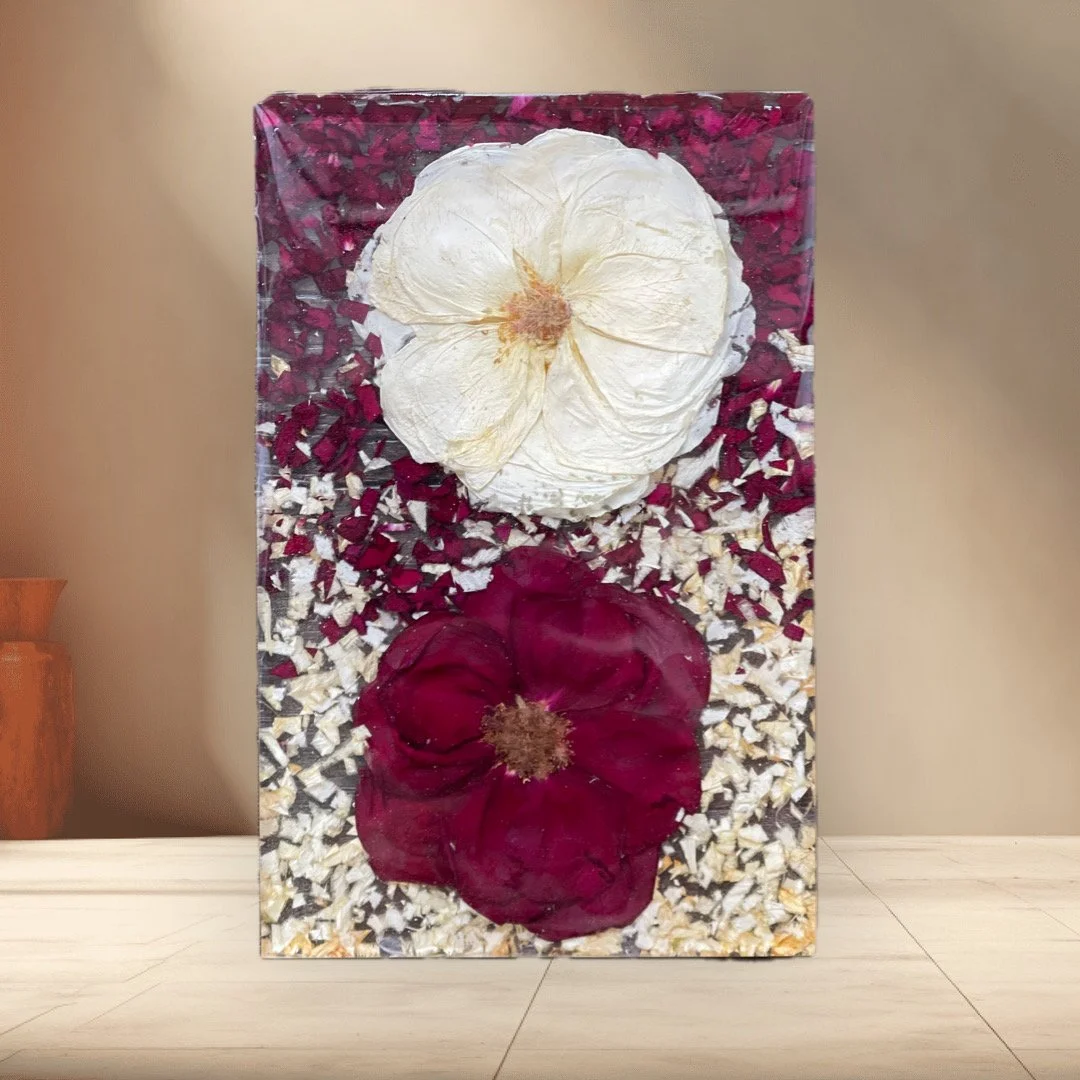 Decorative artwork featuring two large flowers, one white and one dark red, embedded in a rectangular frame with a background of small red, white, and clear fragments.