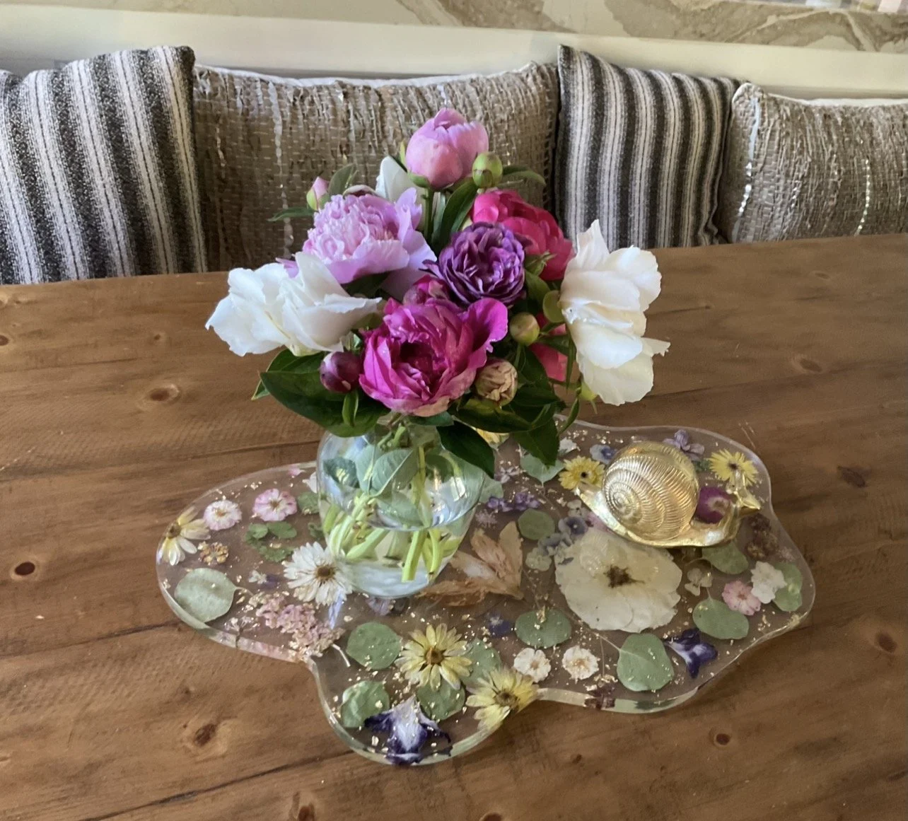 A floral arrangement of pink, white, and purple peonies in a glass vase, placed on a decorative floral tray with a gold snail shell and flower embellishments on a wooden table. In the background, there are striped and textured pillows.