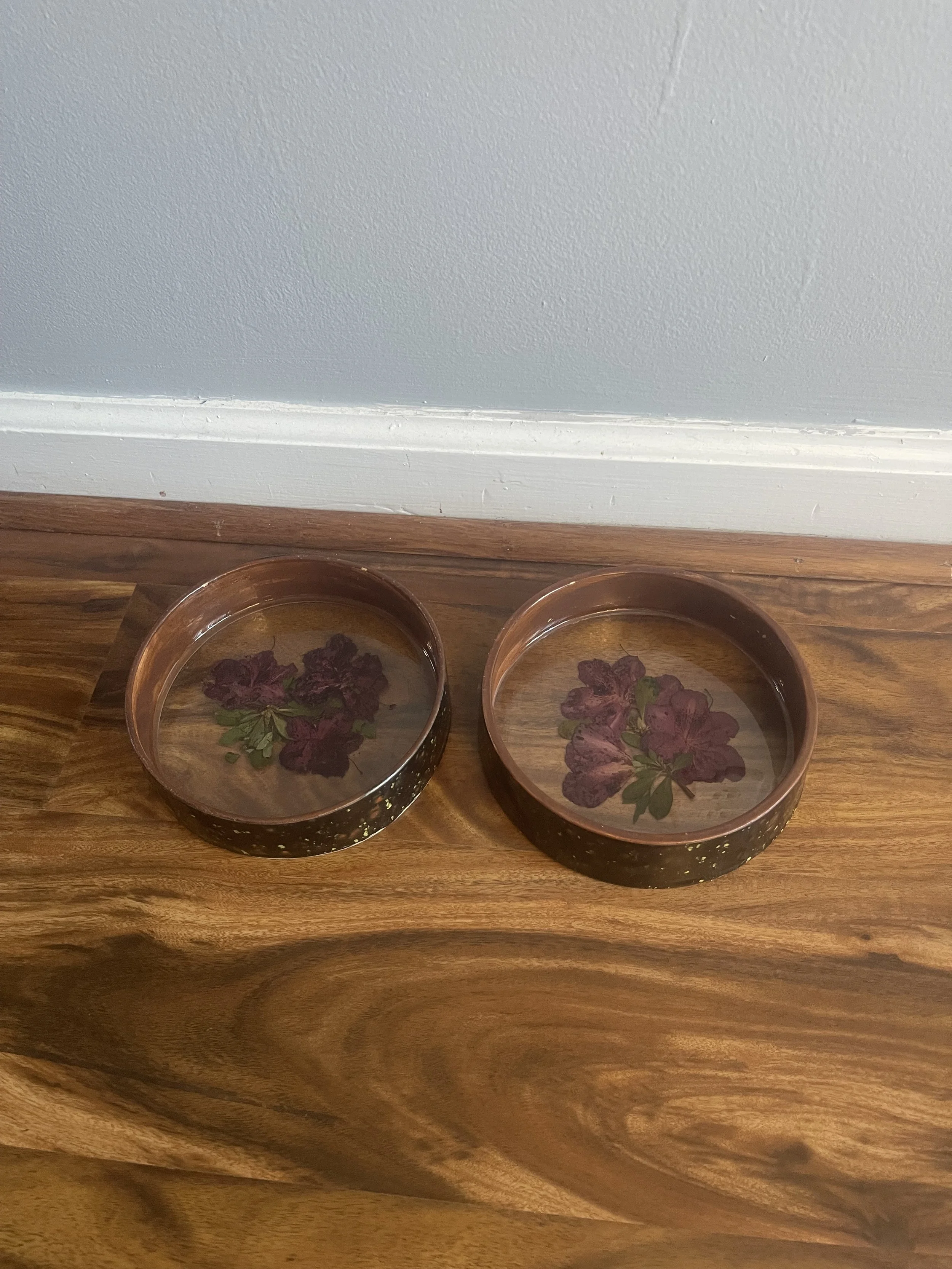 Two brown bowls filled with water, each with a purple flower floating inside, placed on a wooden surface against a light-colored wall.
