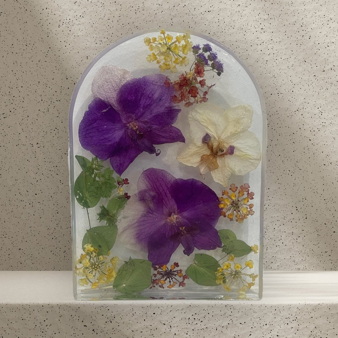 A glass rectangular container filled with various pressed flowers and green leaves, including purple, white, yellow, and orange flowers, placed on a speckled beige surface.