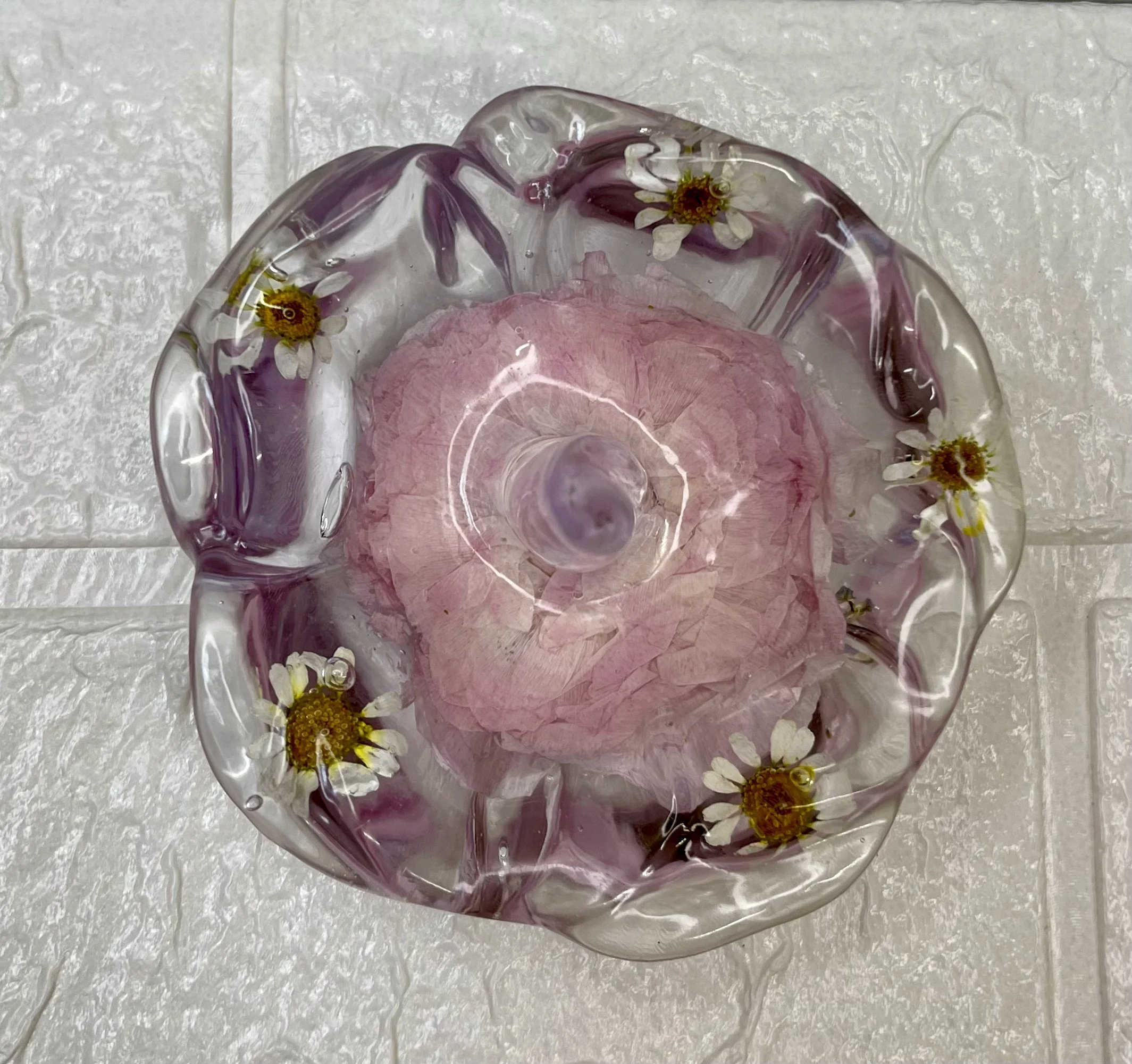 A glass bowl with daisy flowers embedded around the rim, holding a pink tissue paper flower, placed on a textured white surface.