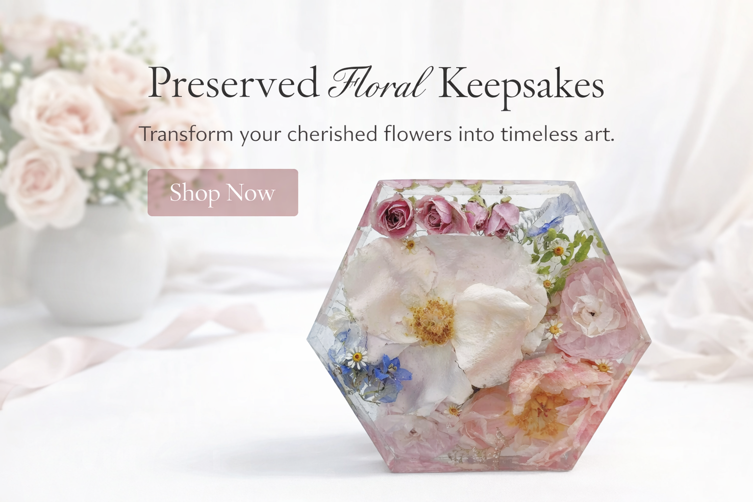 A hexagonal glass display with preserved pink and white flowers, with a blurred pink flower arrangement in a white vase in the background, promoting preserved floral keepsakes with a 'Shop Now' button.
