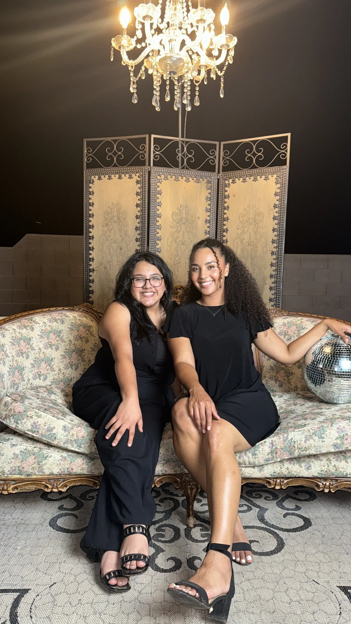 Two women sitting on a vintage floral sofa in a room with a chandelier overhead and a decorative screen behind. One woman has curly hair and the other has straight hair, both are smiling and dressed in black. There is a disco ball on the sofa beside them.