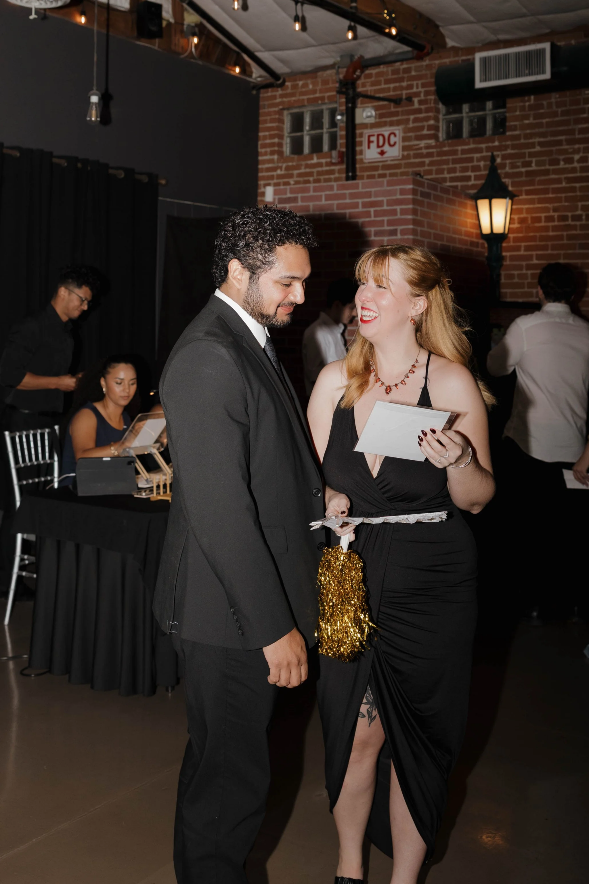 A man and woman dance together at a formal event. The man is wearing a black suit and tie, and the woman is in a black dress with a high slit, red lipstick, and jewelry. They are smiling and holding each other's hands. The background shows other gues