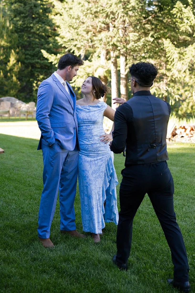 A woman in a light blue dress talking to a tall man in a light blue suit, both standing on grass. A man in black attire with glasses is taking a photo of them against a backdrop of trees.