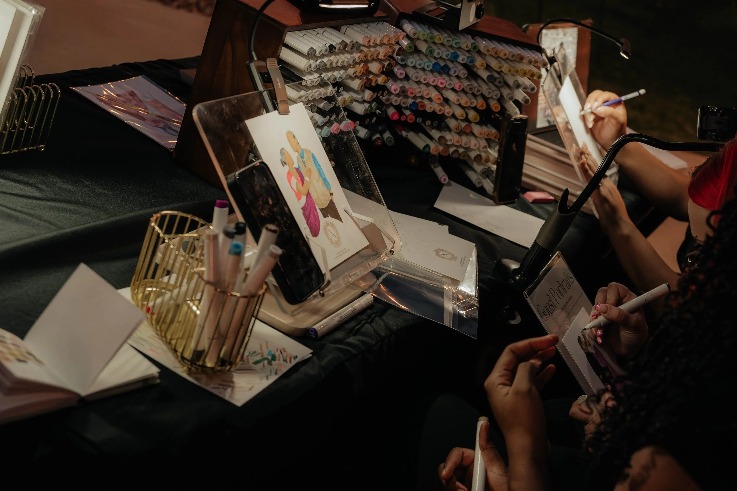 People signing autographs at a table with markers, with a display of markers and artwork on the table.
