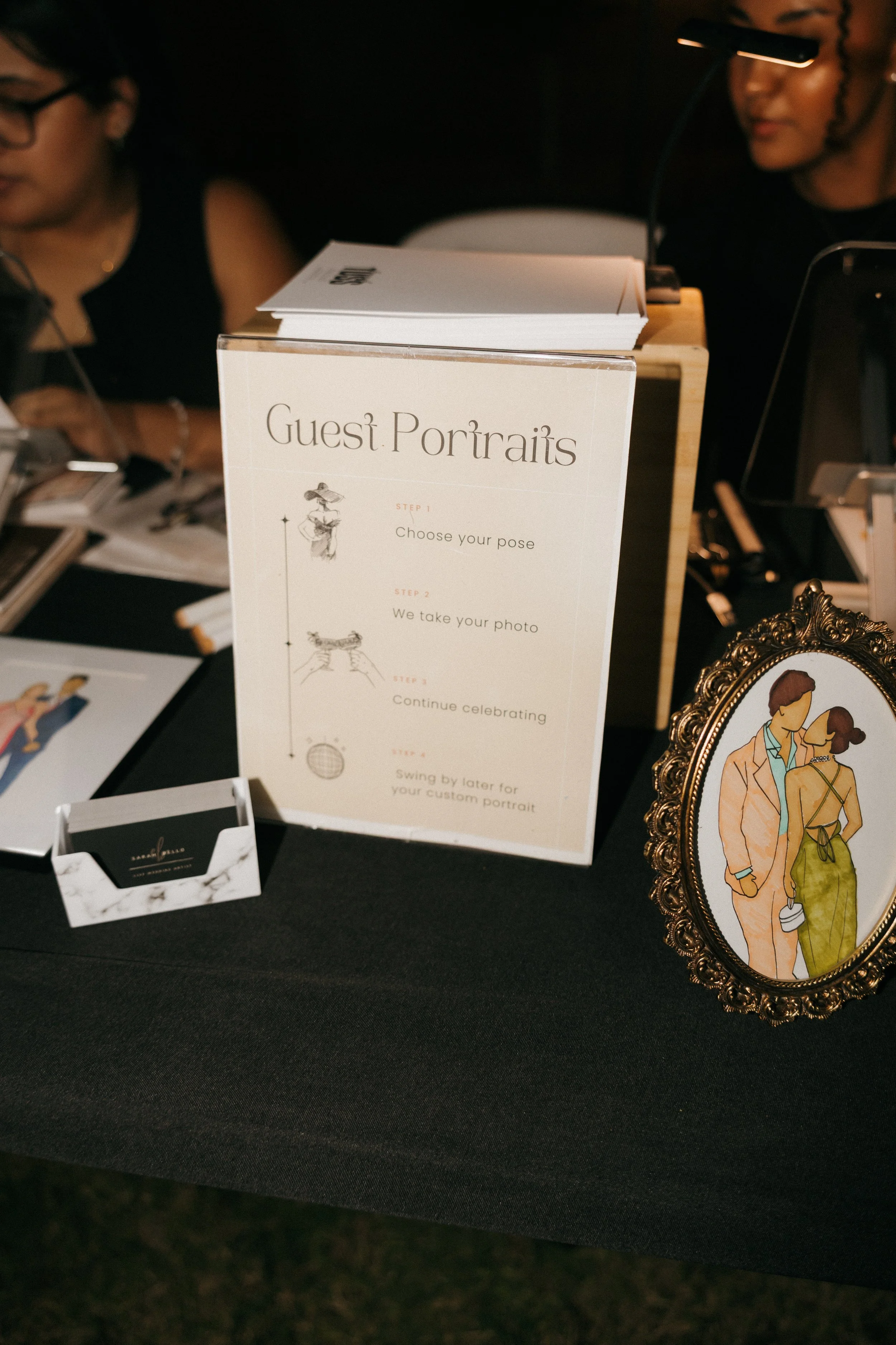 Sign titled 'Guest Portraits' explaining the portrait session steps: choose your pose, we take your photo, continue celebrating, swing by later for your custom portrait. There is a framed illustration of a couple and a small business card or sign on 
