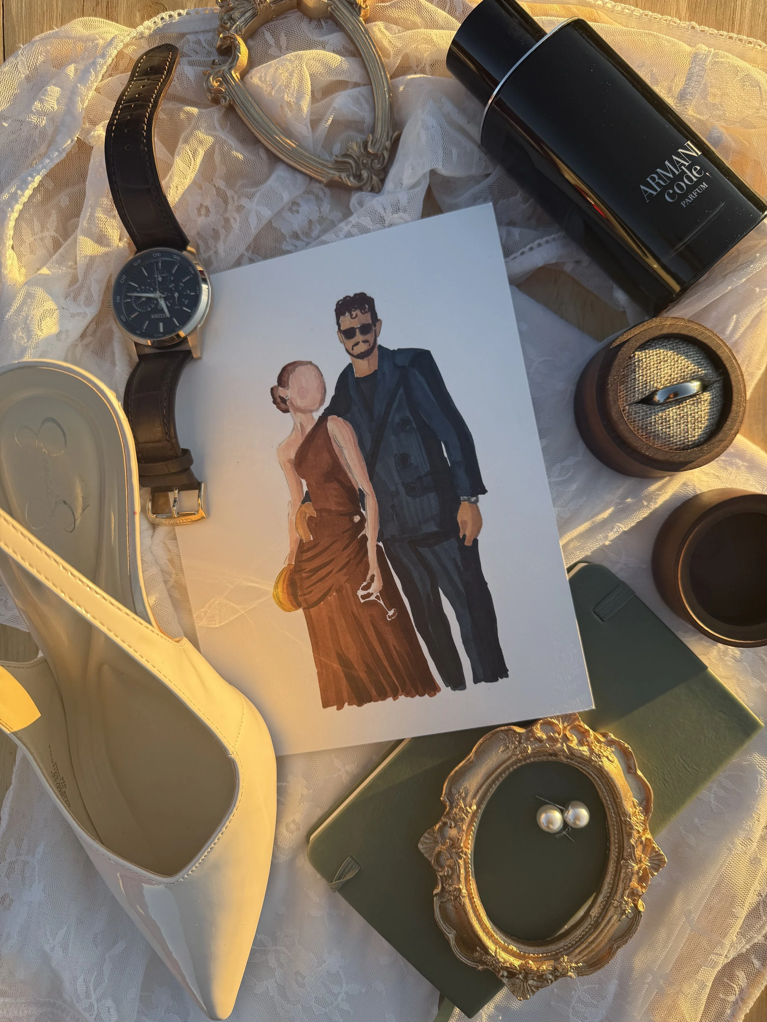A collection of various items on lace fabric, including a wristwatch with a black leather strap and dark dial, a yellow high-heeled shoe, a black can of Armani Code perfume, a gold-framed mirror, an ornate gold picture frame with pearls, a small gree