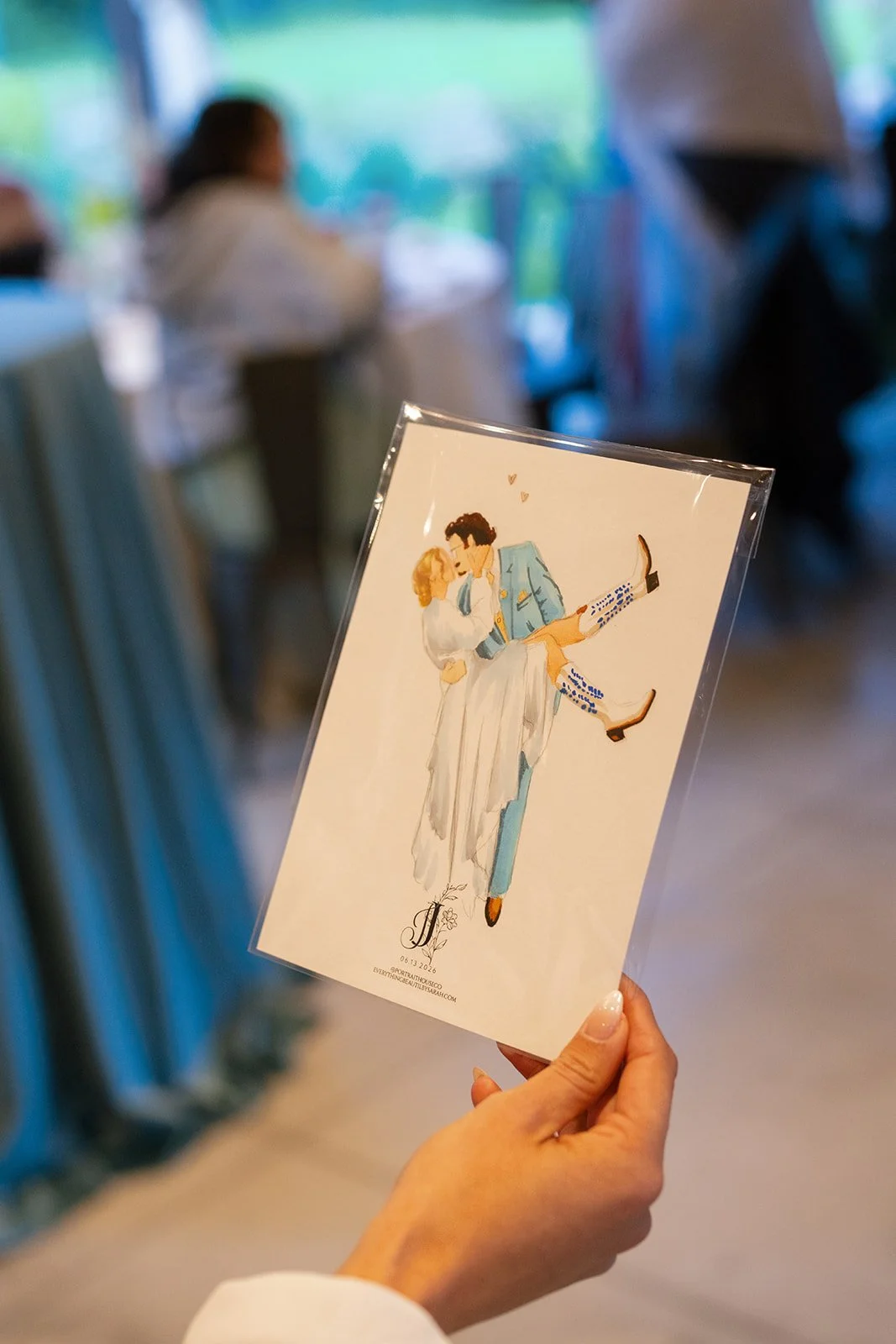 A hand holding a caricature illustration of a couple in wedding attire, with a blurred background of a reception area with people and tables.