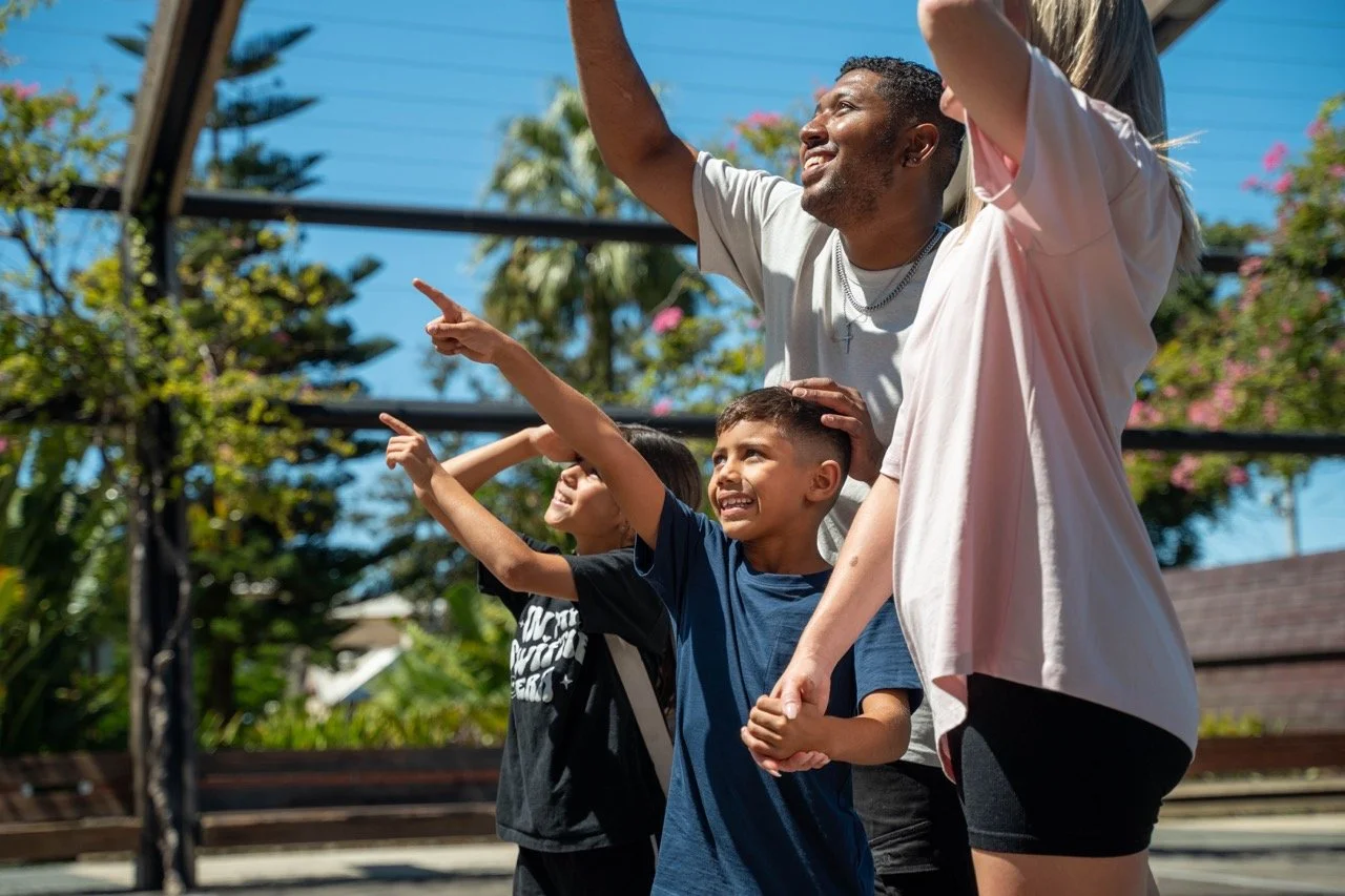 A family of four, including a man, a woman, and two children, smiling and pointing into the sky outdoors on a sunny day.