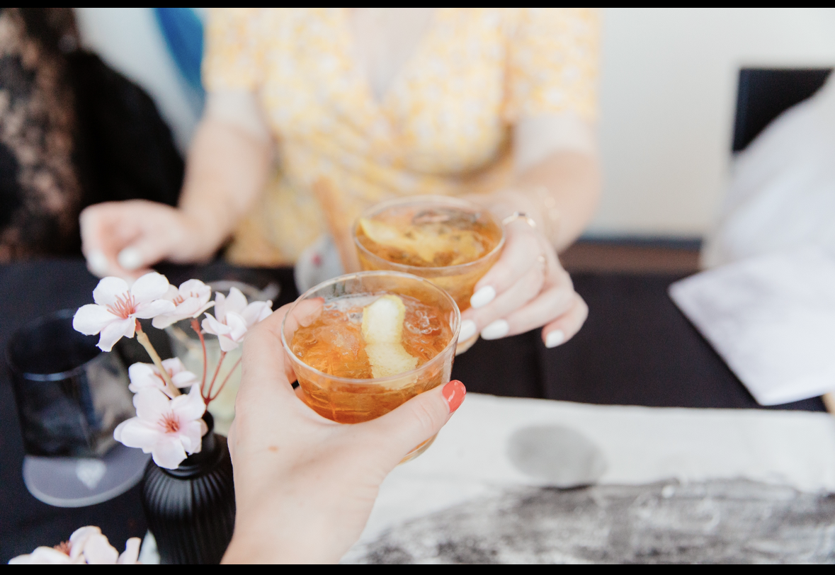 Two people are toasting with glasses of what appears to be a cocktail or wine, with a flower arrangement on a table in the foreground and a blurred background.