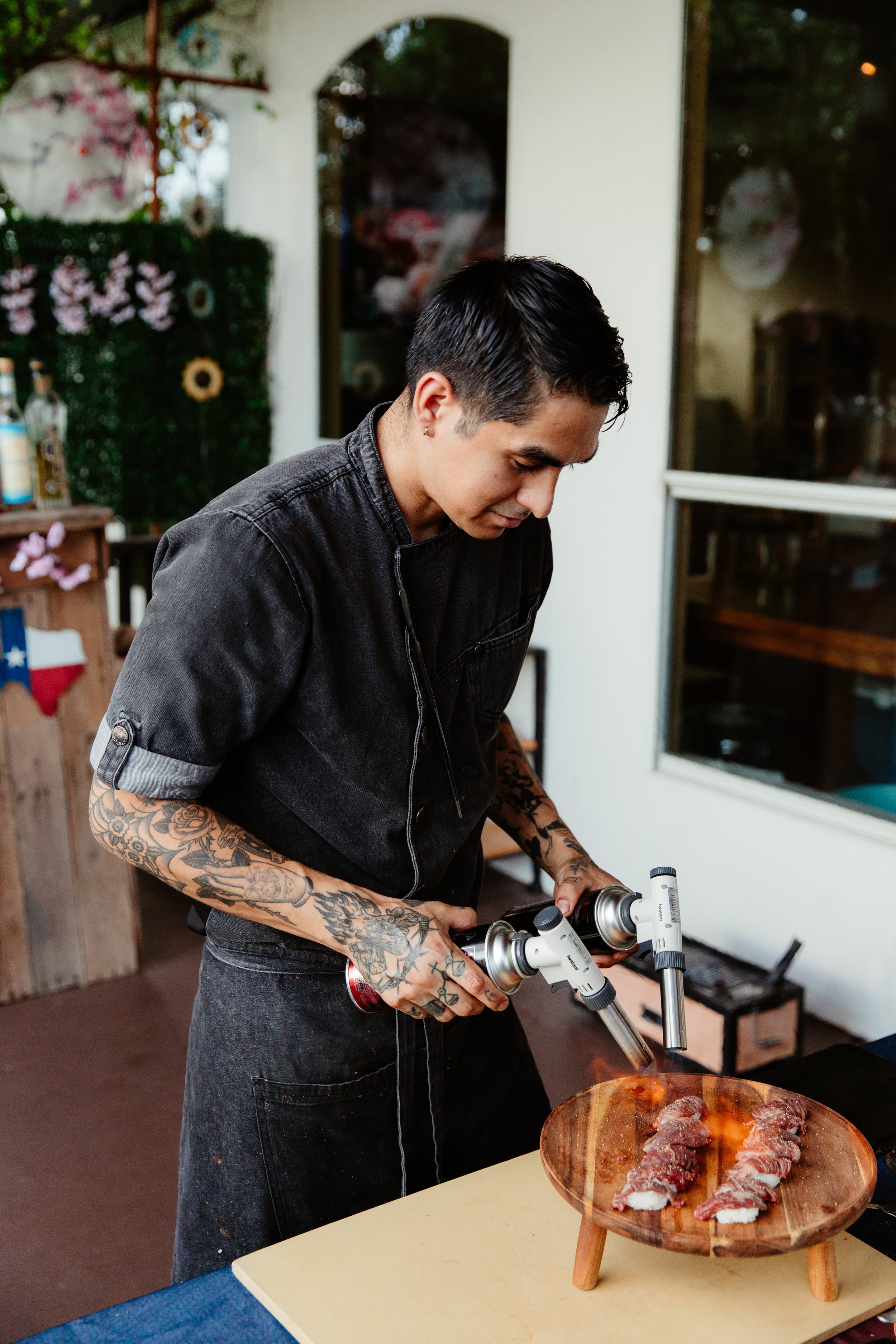 A man with tattoos on his arms and short dark hair, wearing a black denim shirt, is searing meat with a blowtorch over a wooden plate of sliced raw meat, in a home kitchen or restaurant setting.