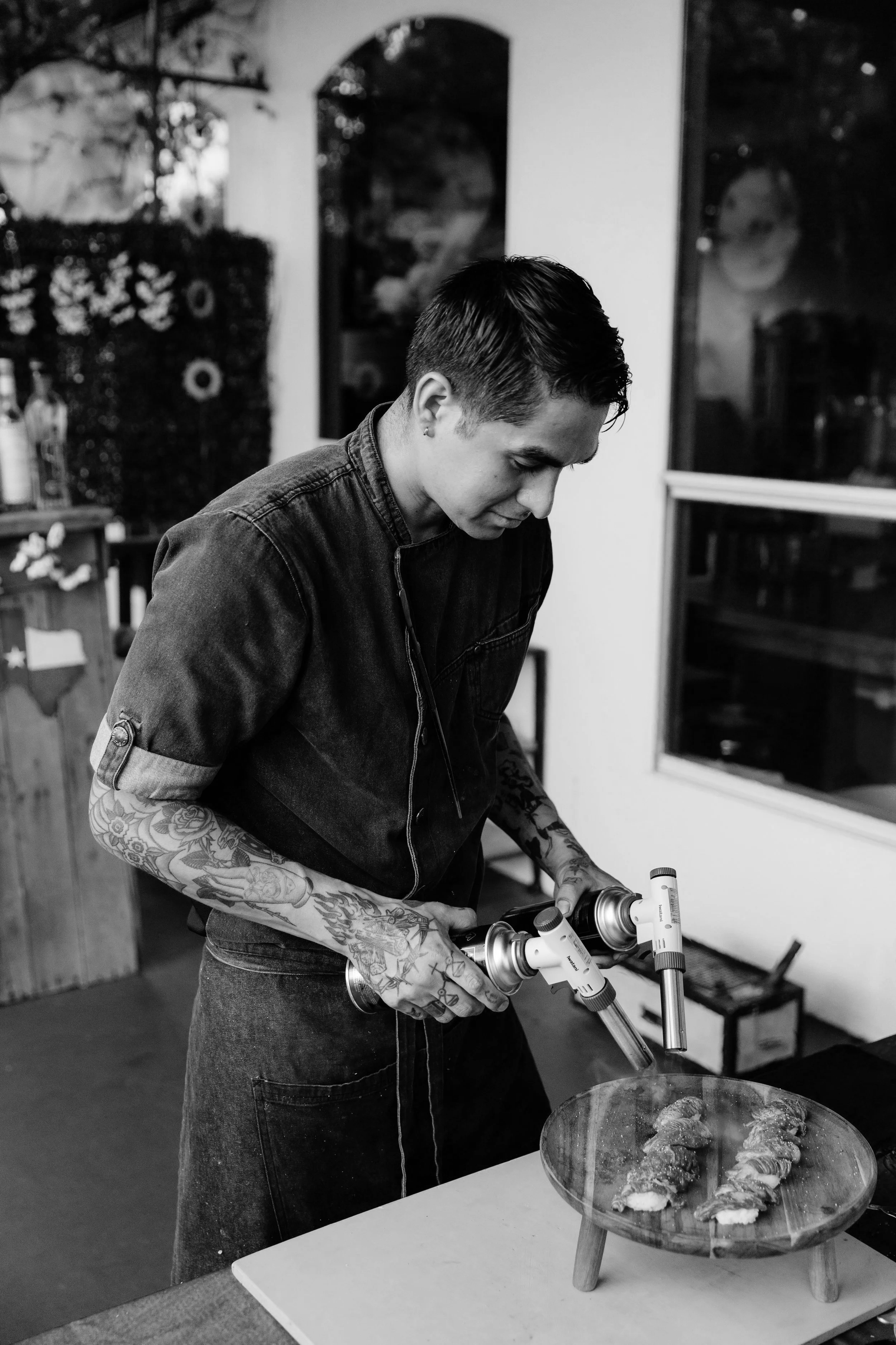A person with tattoos on their arms wearing a denim jacket and jeans, using a spray paint gun to decorate cookies on a glass plate in a kitchen or studio setting.