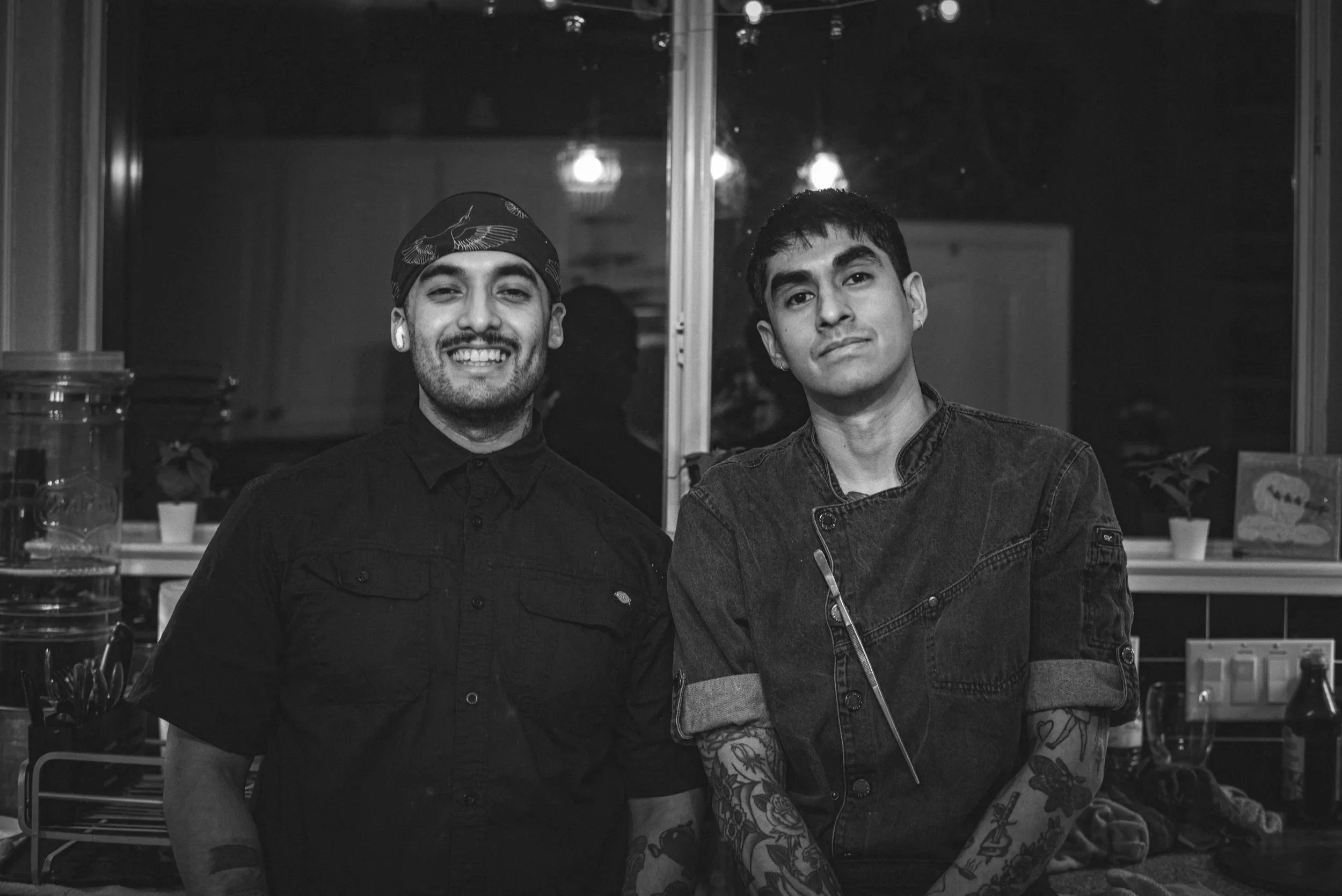 Two young men standing side by side in a room, smiling for the camera. One has short, dark hair and wearing a denim jacket; the other has a headscarf and earbuds, dressed in dark clothing.