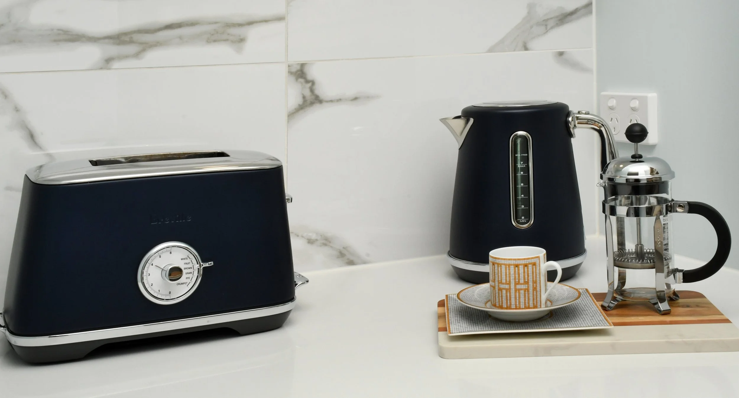Cloudview kitchen vignette featuring grey and white calacatta marble style tiling, a classic navy blue toaster and kettle,  paired with a small gold espresso cup and saucer and a stainless steel French coffee press.