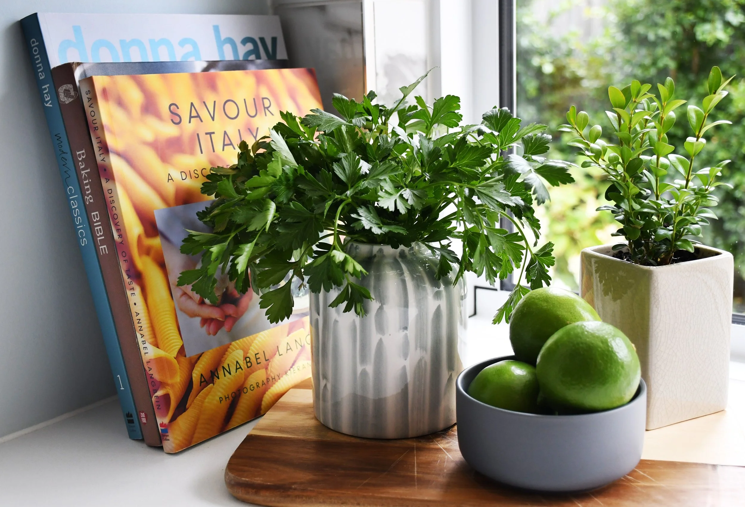 Cloudview kitchen vignette featuring recipe books alongside Italian parsley, green limes and a small green English box each housed in glazed stoneware vessels.