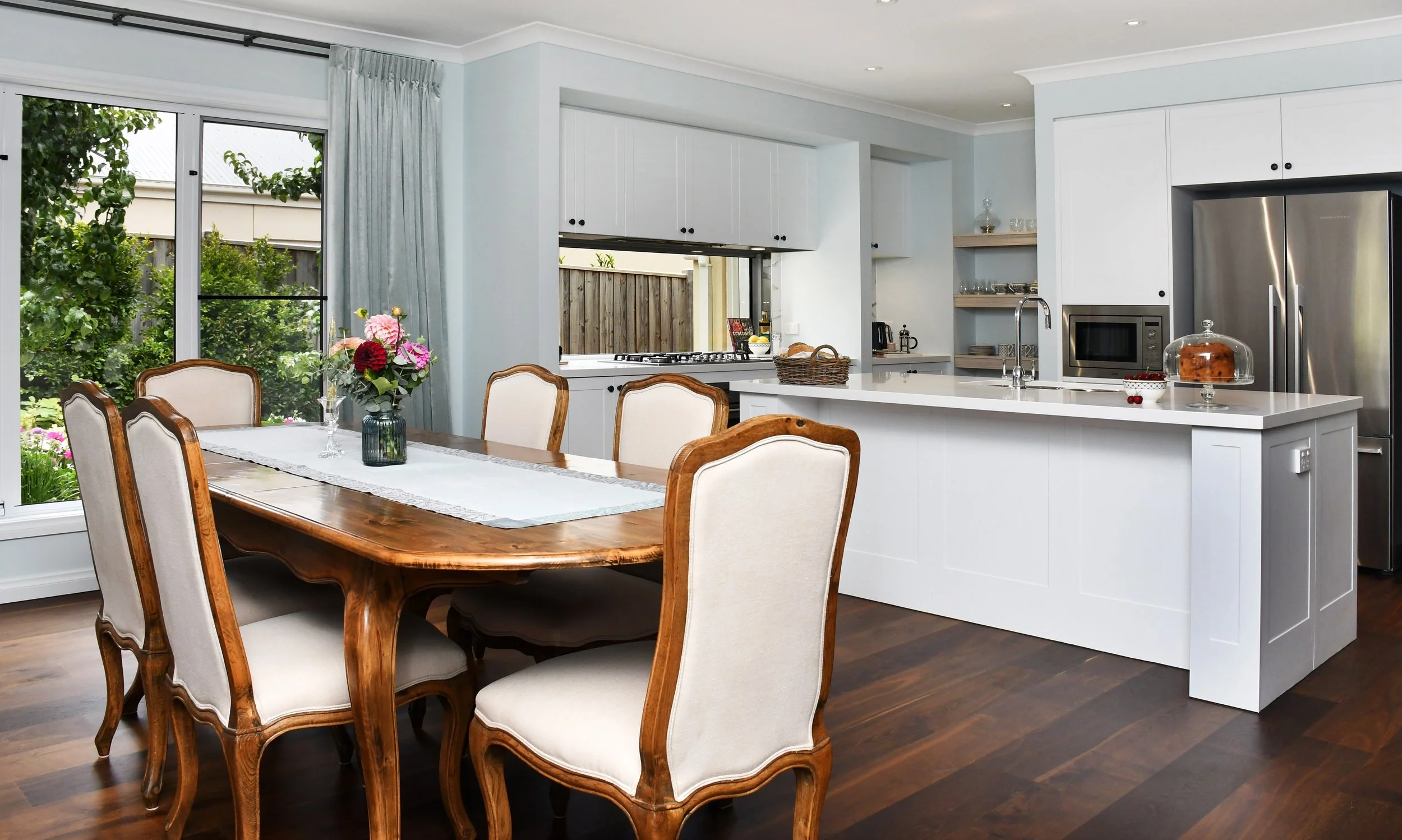 Cloudview dining room looking towards the kitchen featuring French oak Louis XIV style chairs in taupe linen and an oval table with cabriole legs. The lovely east garden and window boxes frame the background and flood the space with morning light.