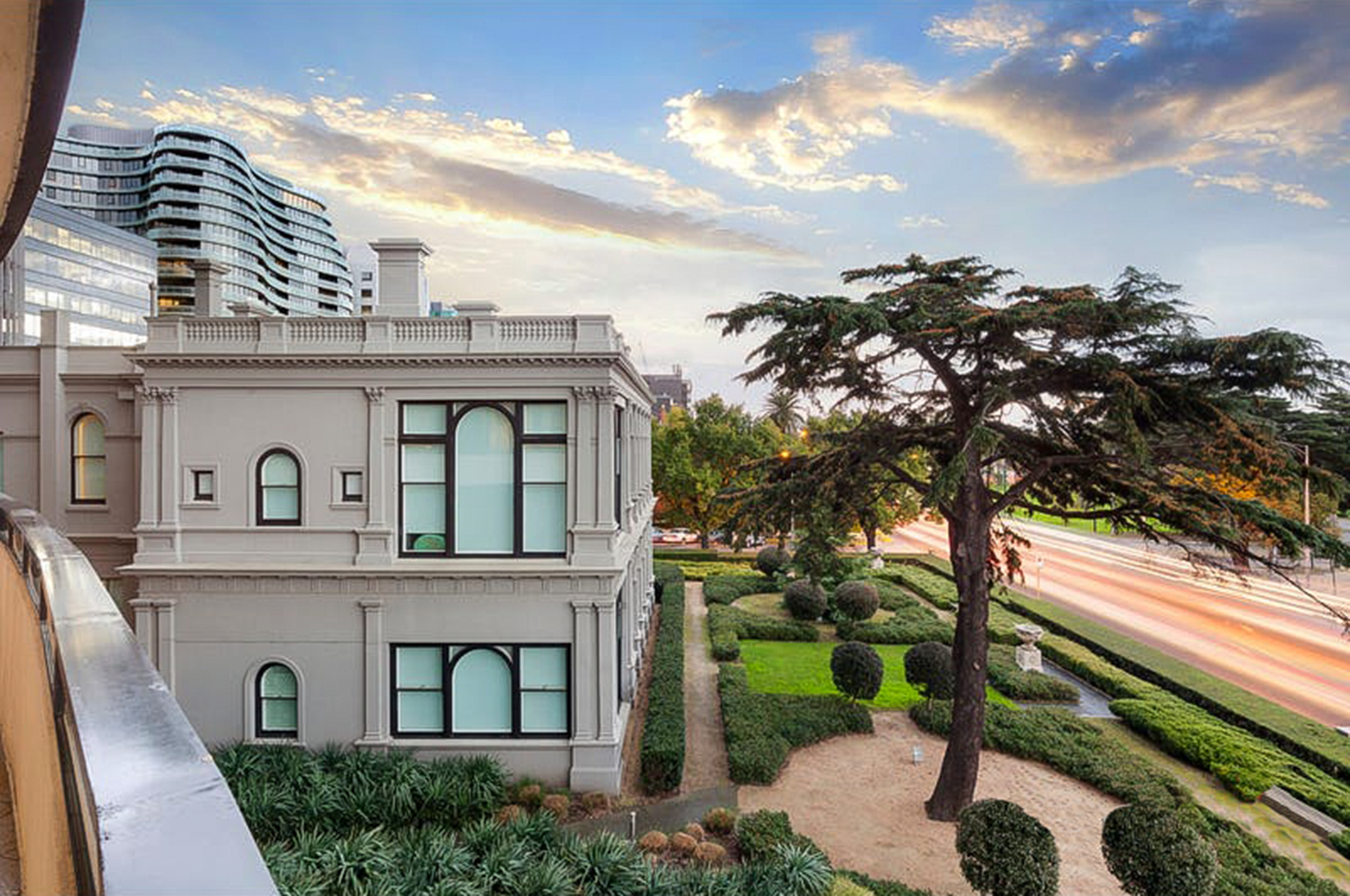 The grand facade of a white Victorian mansion, viewed from the Parklife Apartment balcony, is offset by the old cedar tree and deep evergreens of the English style garden in the foreground.
