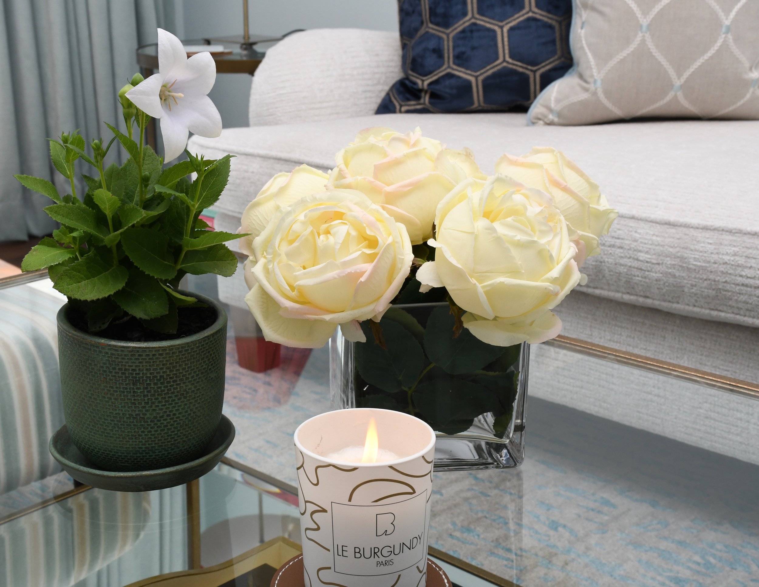 Cloudview snug coffee table vignette with white garden roses in a glass vase, a white lily in a green ceramic pot and a fragrant gold accented candle from Paris. In the background feather down pillows rest on the comfy sofa in shades of navy velvet a