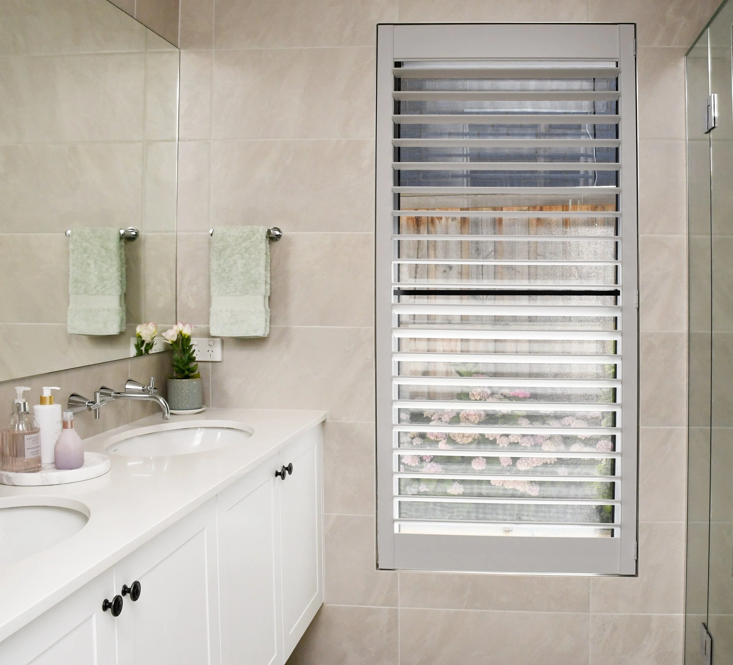 Cloudview ensuite bathroom featuring full height taupe stone tiling, a white shaker style vanity with under mount sinks, large mirror and glass shower recess looking towards the window with white shutters.