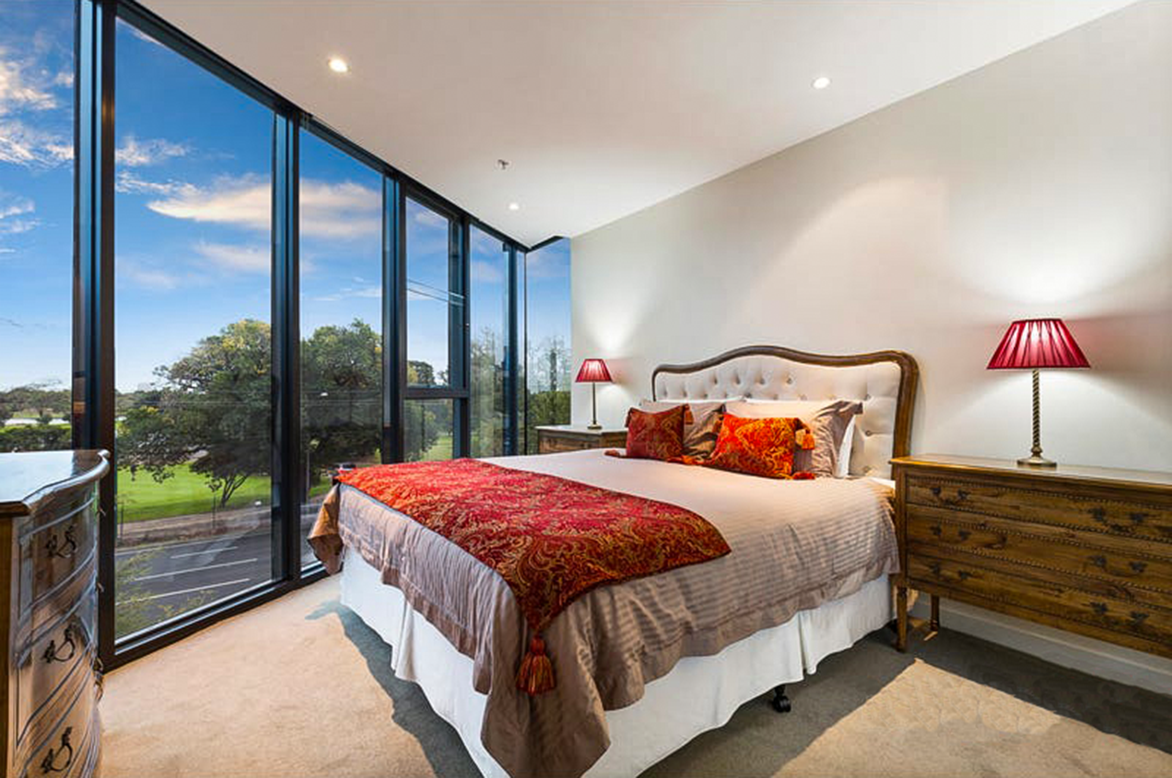 The Parklife Apartment master bedroom overlooks Albert Park through stunning full height windows. The Louis XIV style oak furniture suite elegantly rests in the space with light walls and cosy carpet. A pop of red on the bed side lampshades, cushions