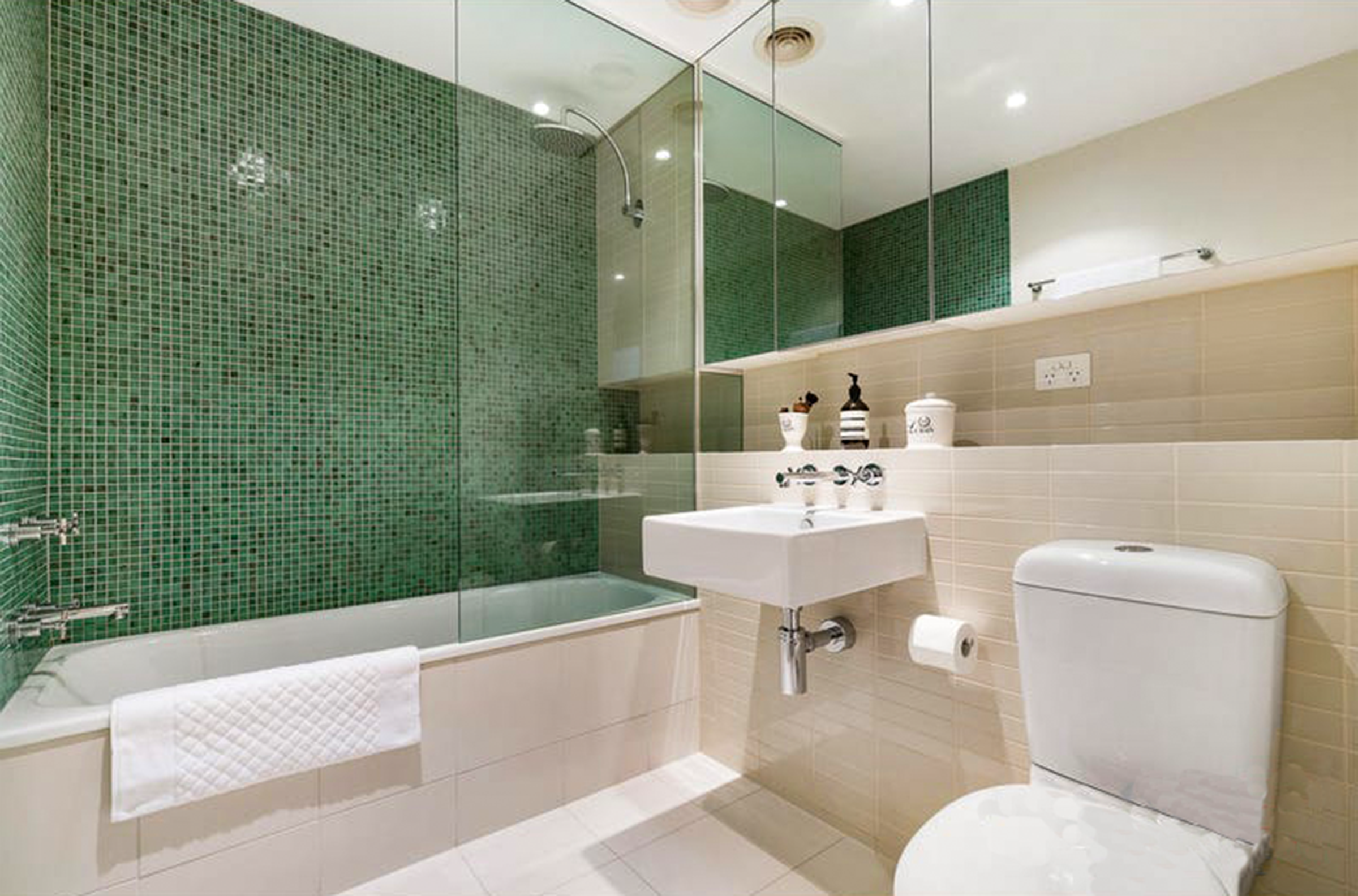 The Parklife Apartment bathroom feels like a serene spa and features off white tiling with a stunning mosaic green feature wall in the combined bath-shower recess. A luxurious rain shower, modern white fixtures and fluffy white towels complete the pi