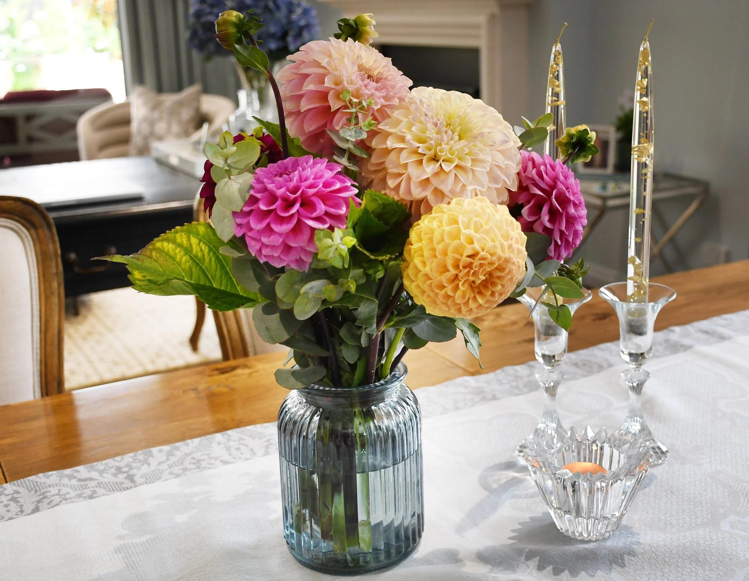 Cloudview interior dining vignette featuring a French style oak dining setting, a duck egg blue table runner, a blue glass vase with dahlia flowers  in pink, apricot and yellow alongside three classic crystal candlesticks.