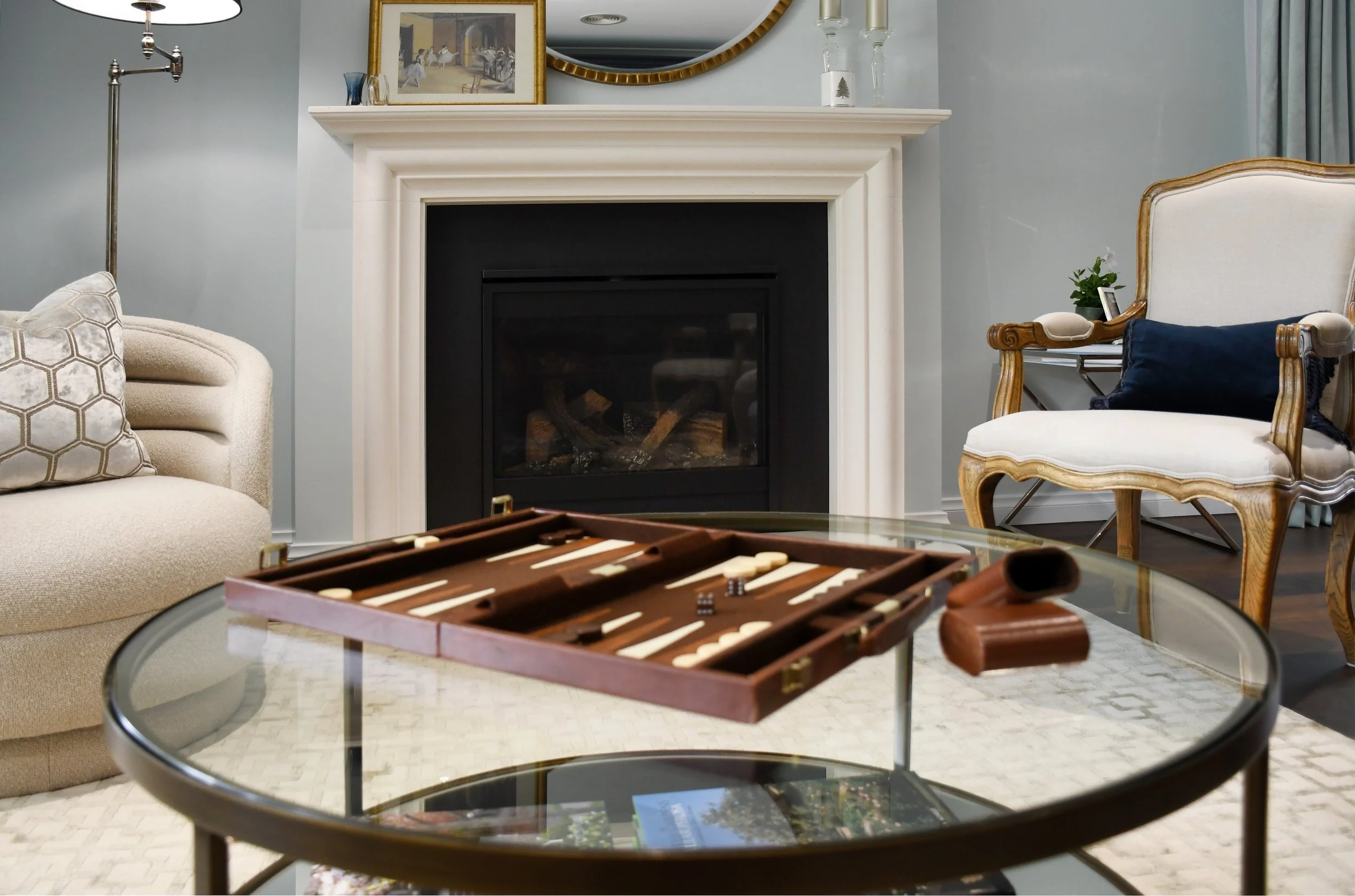 A beautiful living room with a round glass coffee table and a brown and white leather backgammon game laid out ready to play, a cream upholstered tub chair and taupe Louis XIV armchair sit beside the limestone fire place and the coffee table
