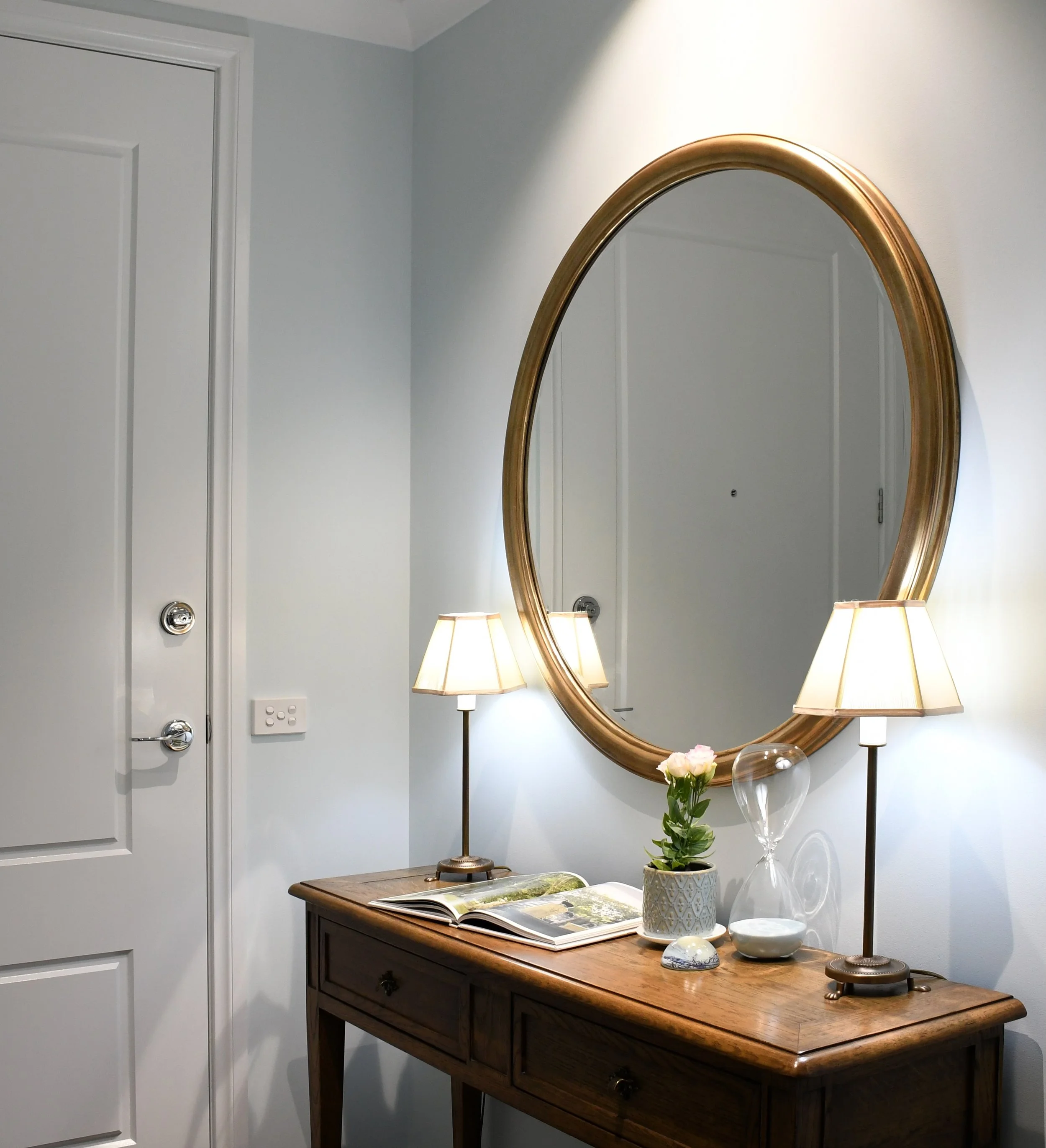 Cloudview entry with front door featuring an oak entry table, large round gold framed mirror, antique brass lamps with cream silk shades and a vignette with a pink lisianthus flower,  an hourglass and an open book.
