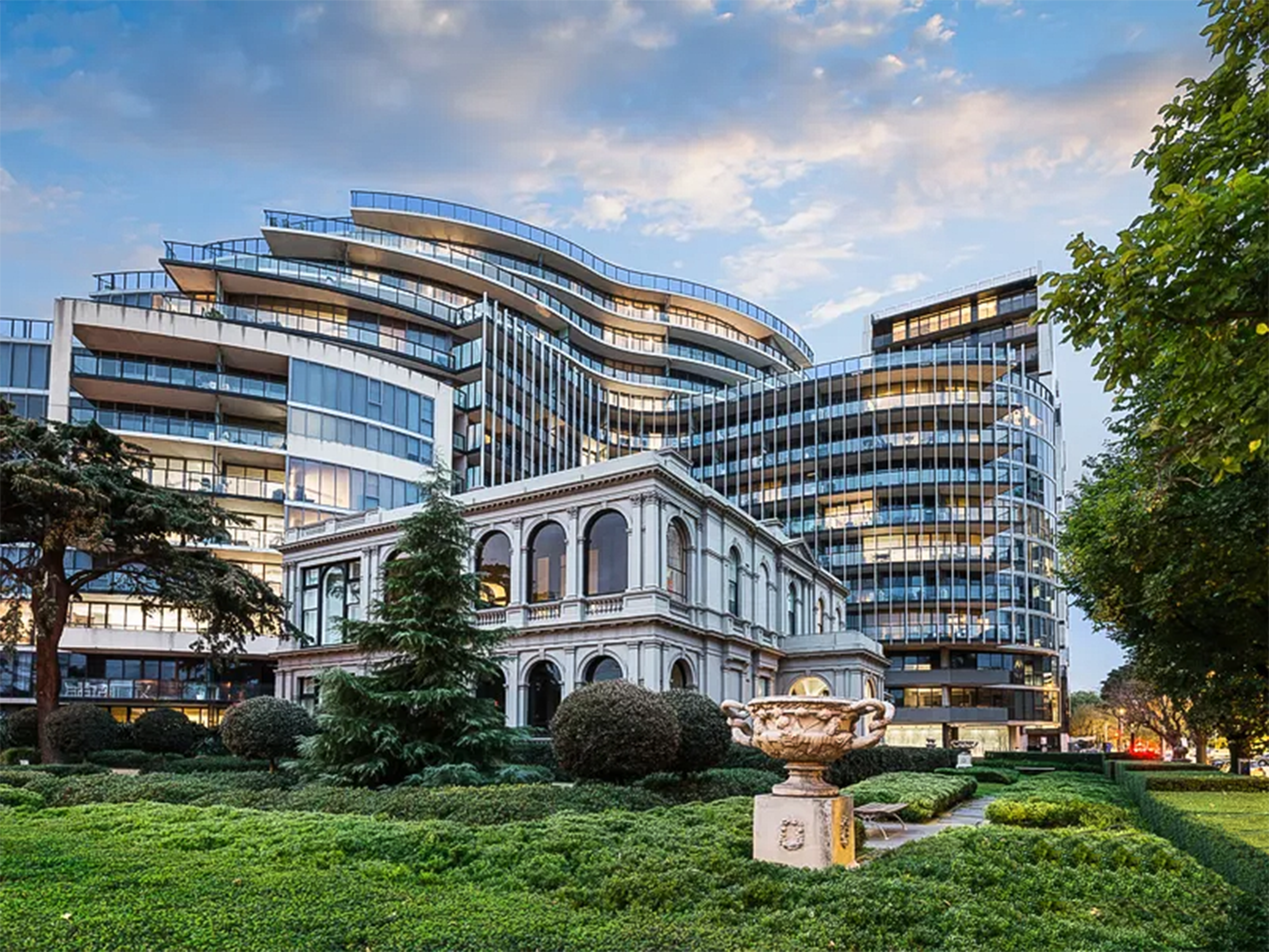 The grand facade of a white Victorian mansion is enveloped in a glass and steel apartment building, which is softened by the classical urns and deep evergreens of the English style garden in the foreground.