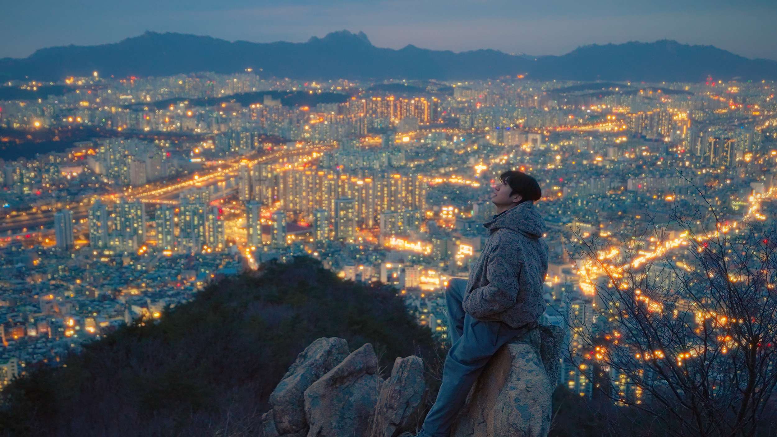 A person sitting on a rock overlooking a cityscape at dusk, with various lights illuminating buildings and streets, and mountains visible in the background.