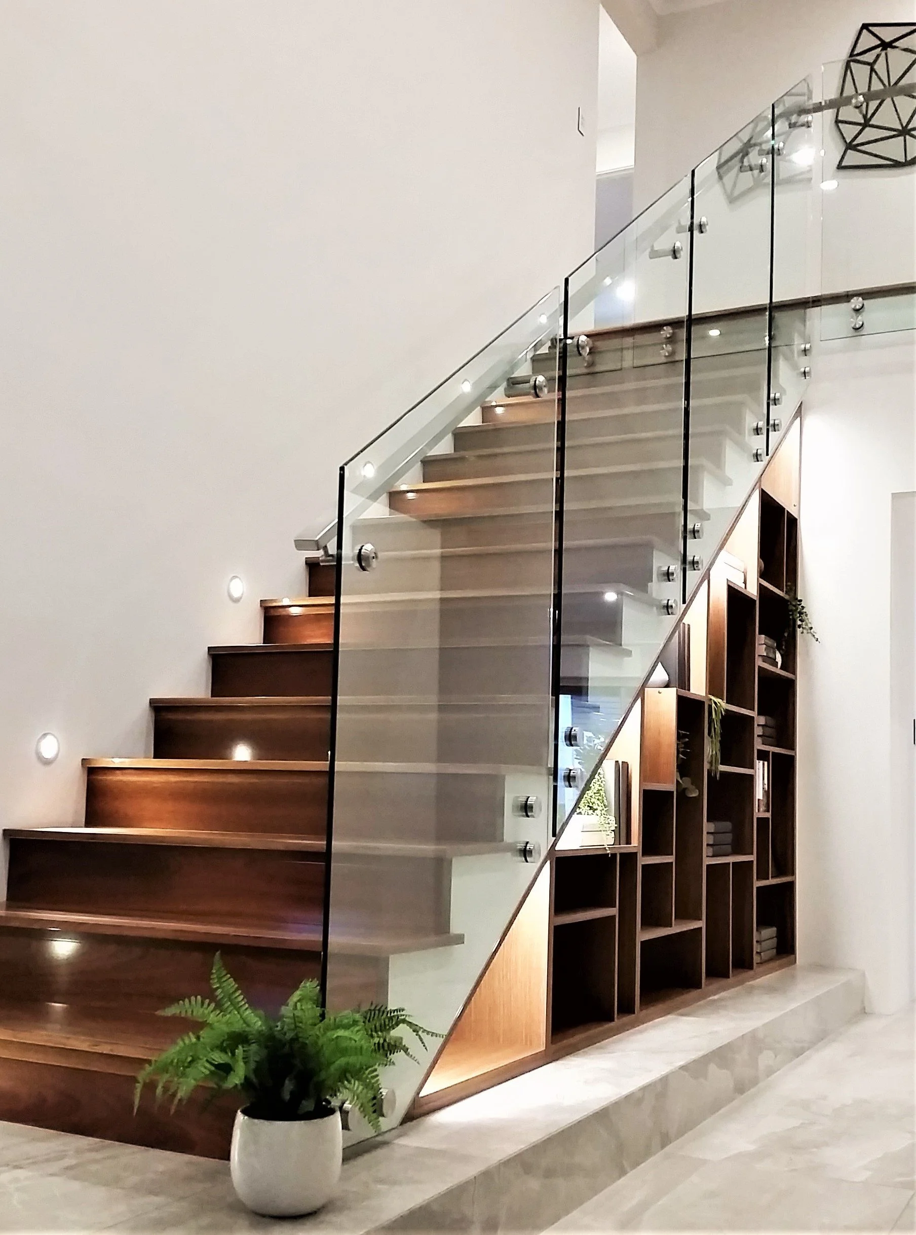 Interior staircase with wooden steps, glass railing, and built-in wooden shelving unit with decorative items and books beneath the stairs, with a potted fern in the foreground.