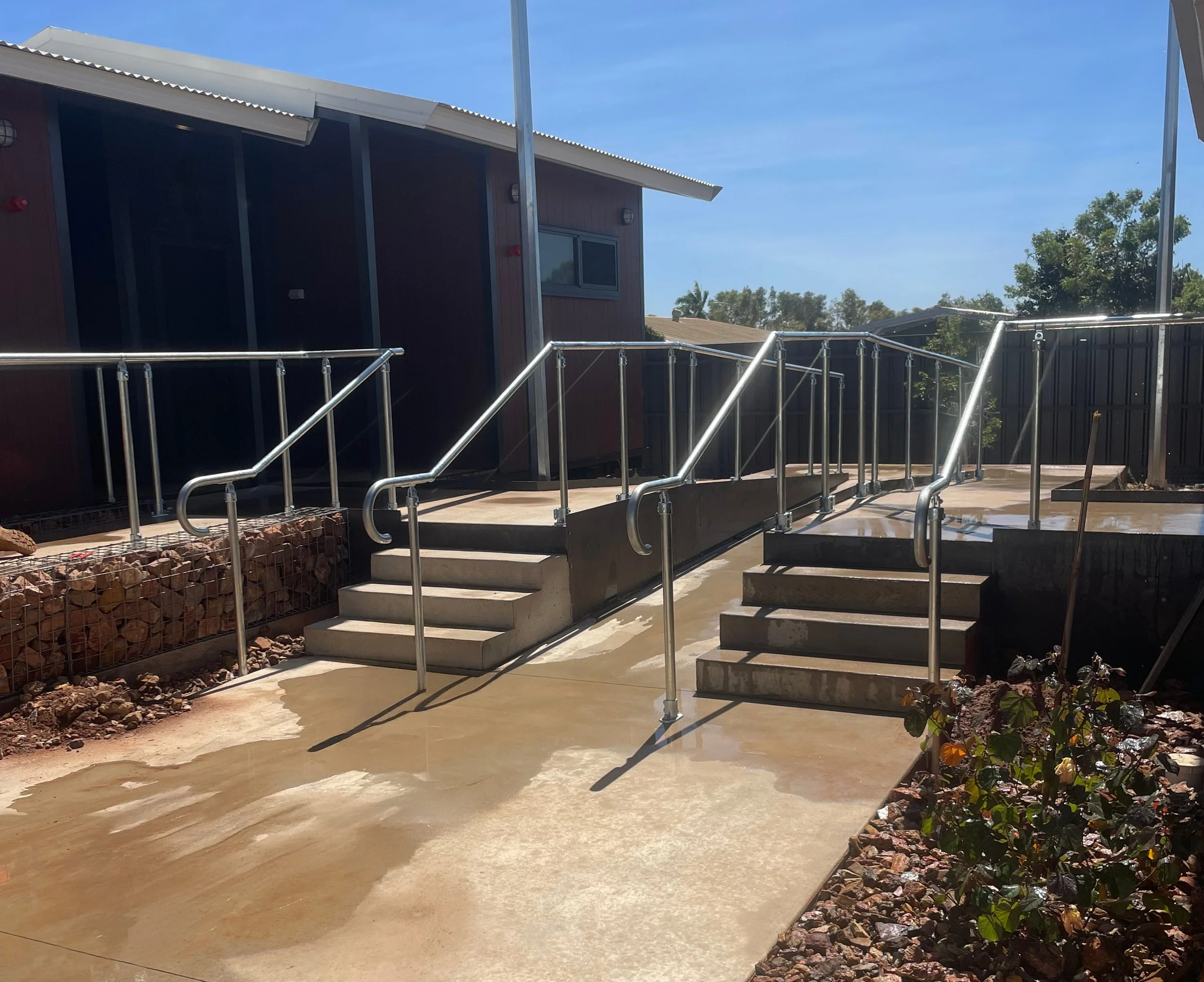 Concrete wheelchair ramp with metal handrails leading to a house entrance, surrounded by landscaping and trees under a clear blue sky.