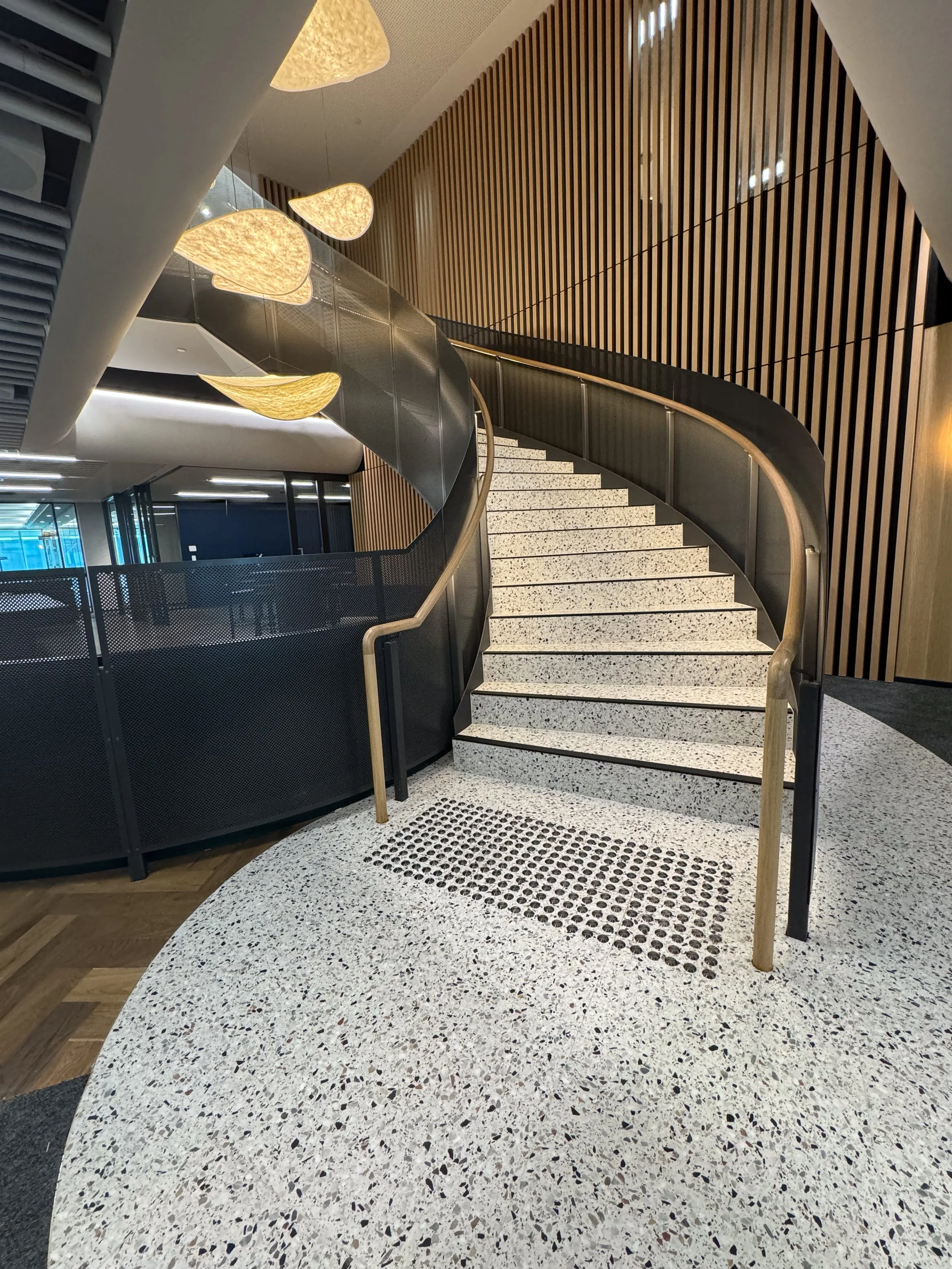 Interior of a modern building with a curved staircase with terrazzo steps, wooden handrails, black metal railing, and wooden wall paneling, featuring artistic hanging light fixtures.