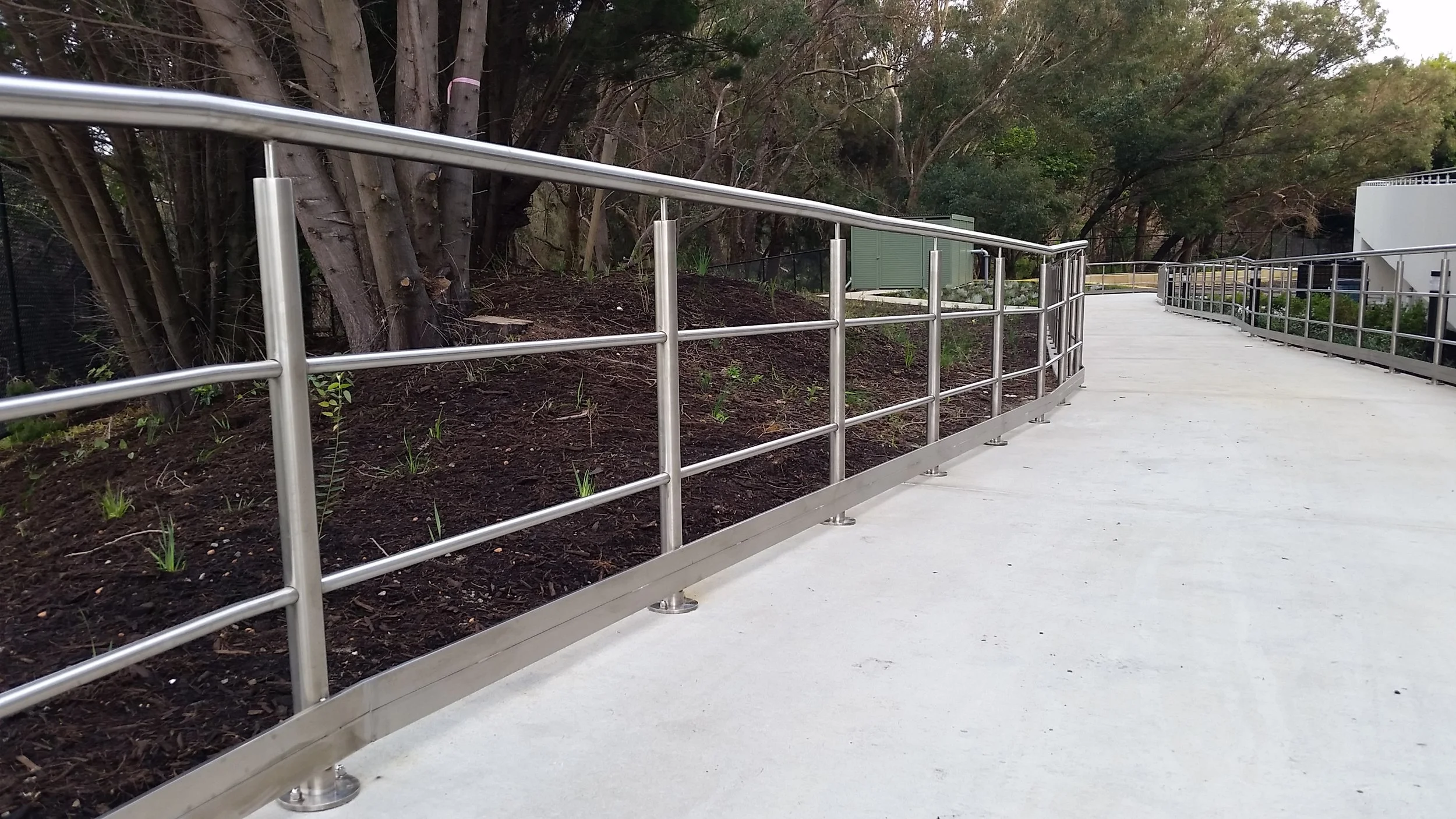 Concrete pathway with metal handrails alongside a garden bed with new plants, trees and bushes in background.