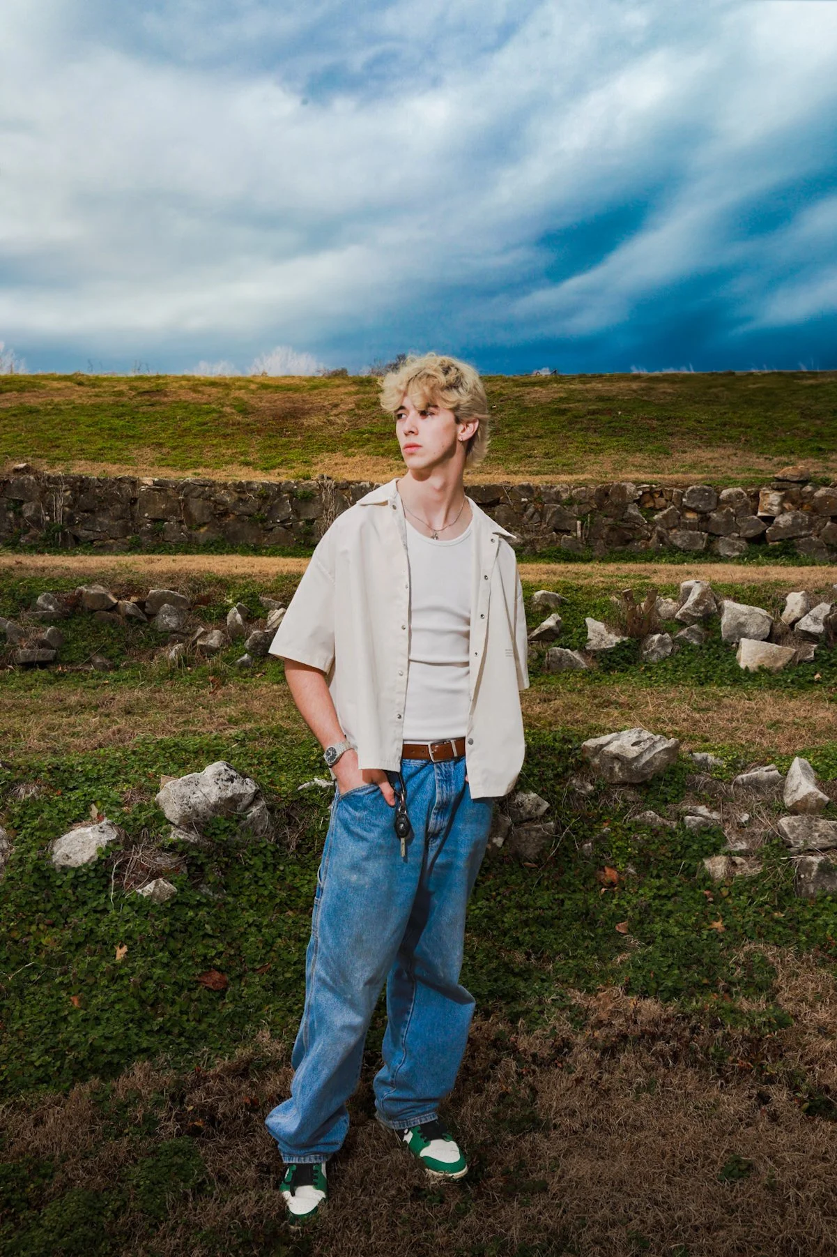 Young man with blond, curly hair standing outdoors on a grassy area with an overcast sky in the background. He is wearing a beige button-up shirt over a white t-shirt, baggy blue jeans, and sneakers. He has a watch and a necklace, and he is holding the keys in his right pocket.