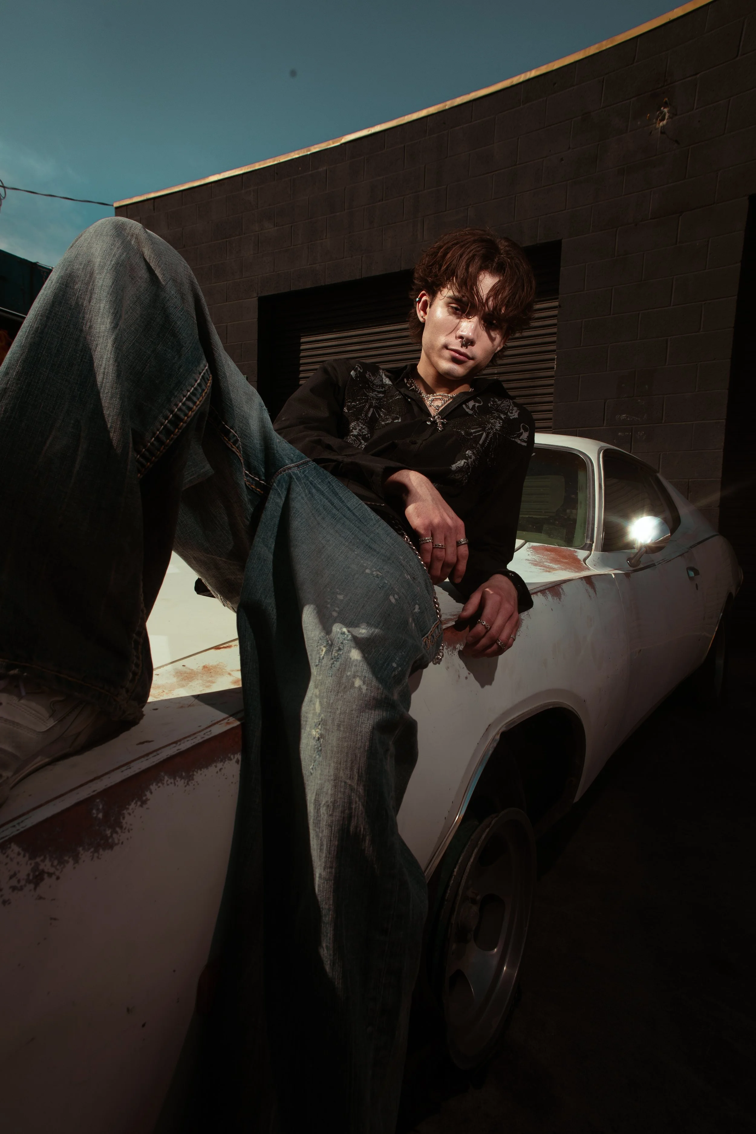 Young man with dark hair, wearing a black shirt and jeans, sitting on the hood of an old white car with rust and dirt, posing in front of a dark brick building under a blue sky.