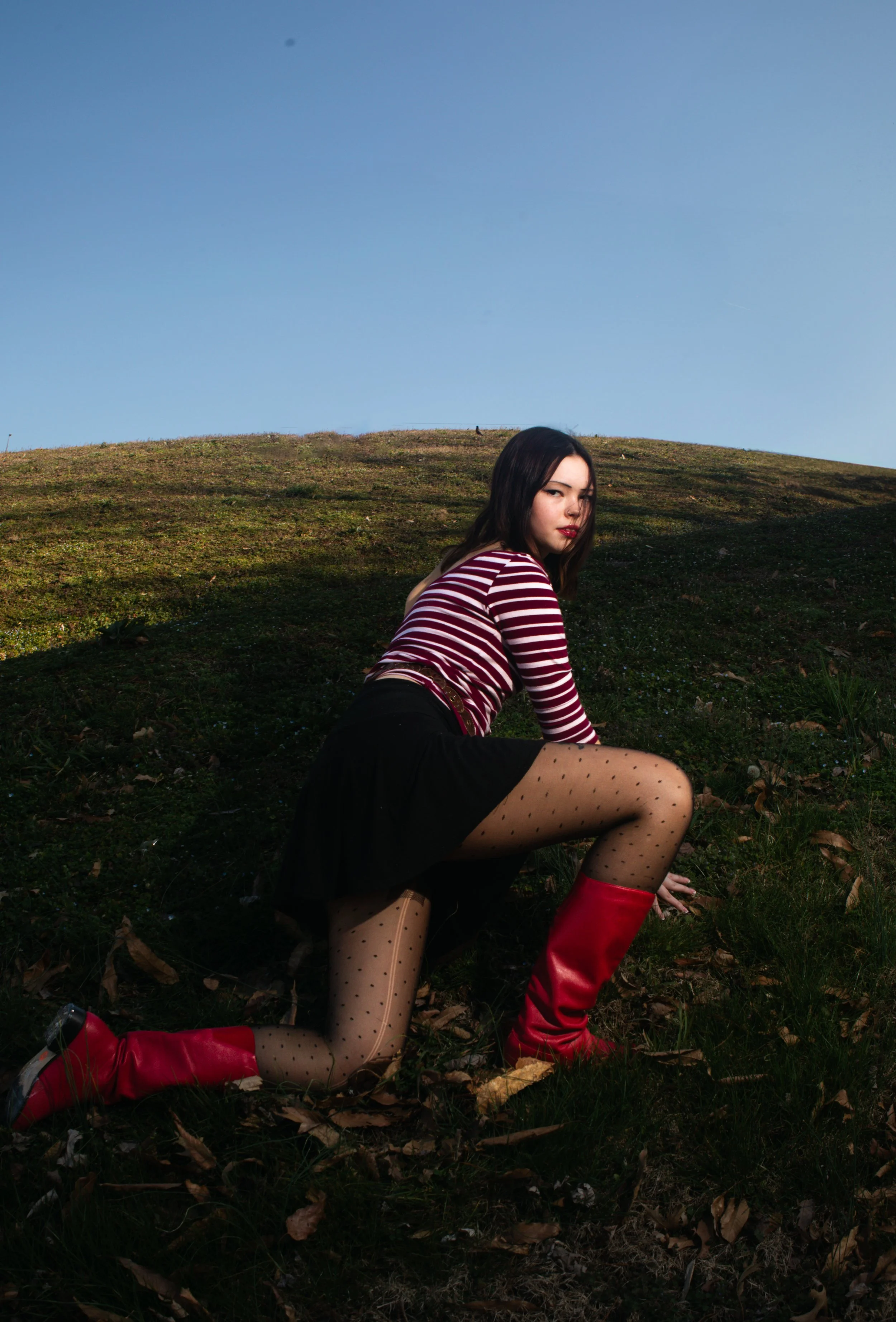 A young woman with dark hair, wearing a red and white striped top, black skirt, polka-dot tights, and red boots, poses on a grassy hillside under a blue sky.