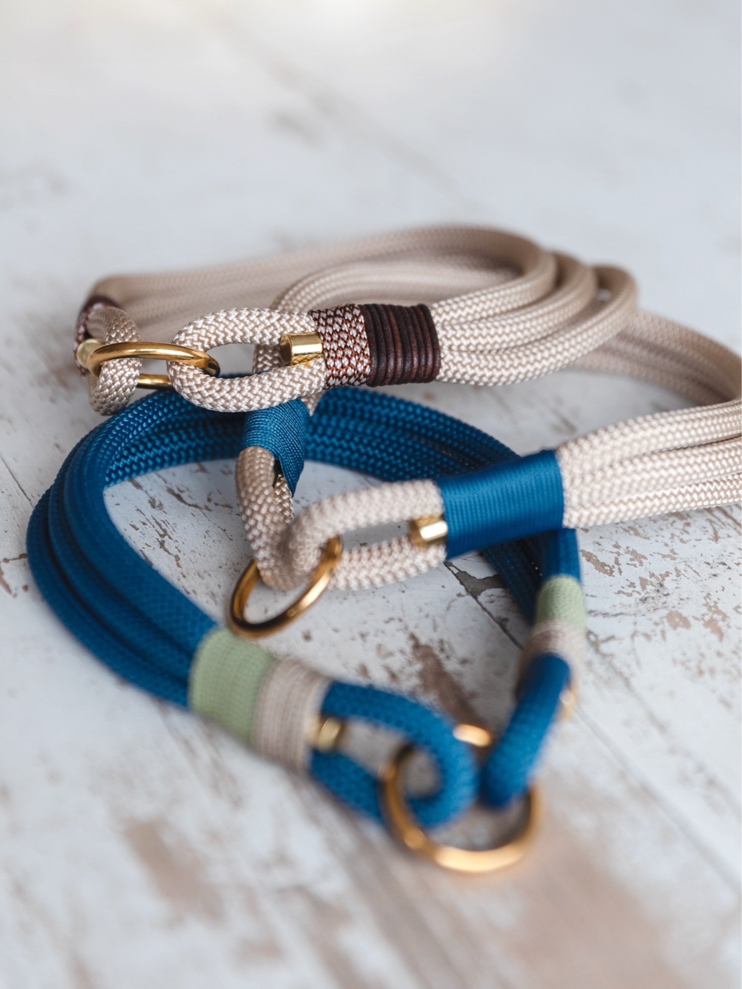 Zwei Hundeleinen aus dickem gewebtem Material in Blau und Beige mit goldenen Haken und D-Ringen auf einem weißen Holzhintergrund.