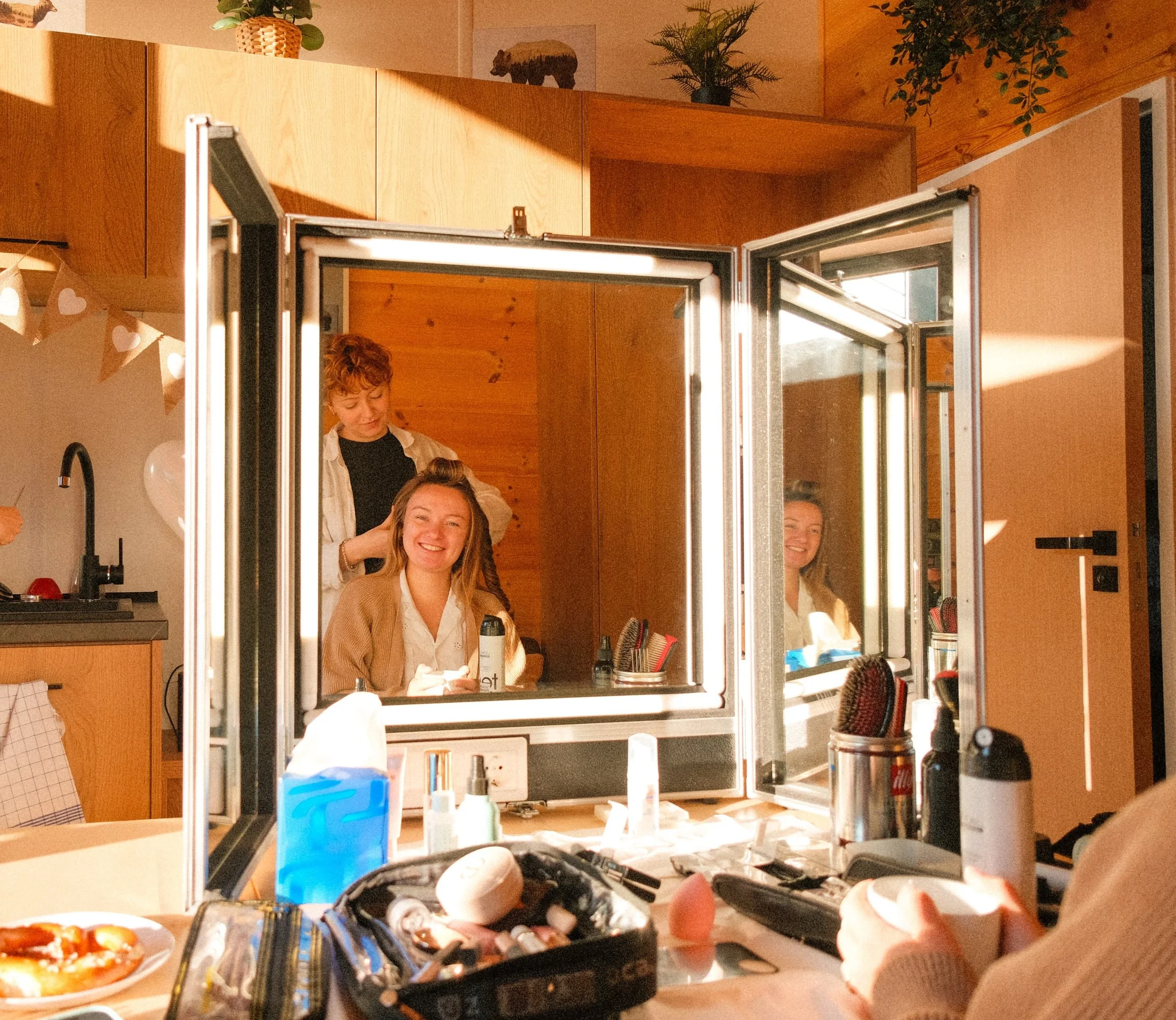 Eine junge Frau sitzt vor einem Spiegel in einem Tiny House in der Glockenalm, während eine Maskenbildnerin ihr die Haare frisiert. Die Szene ist warm beleuchtet und im Hintergrund sind Holzaccessoires.